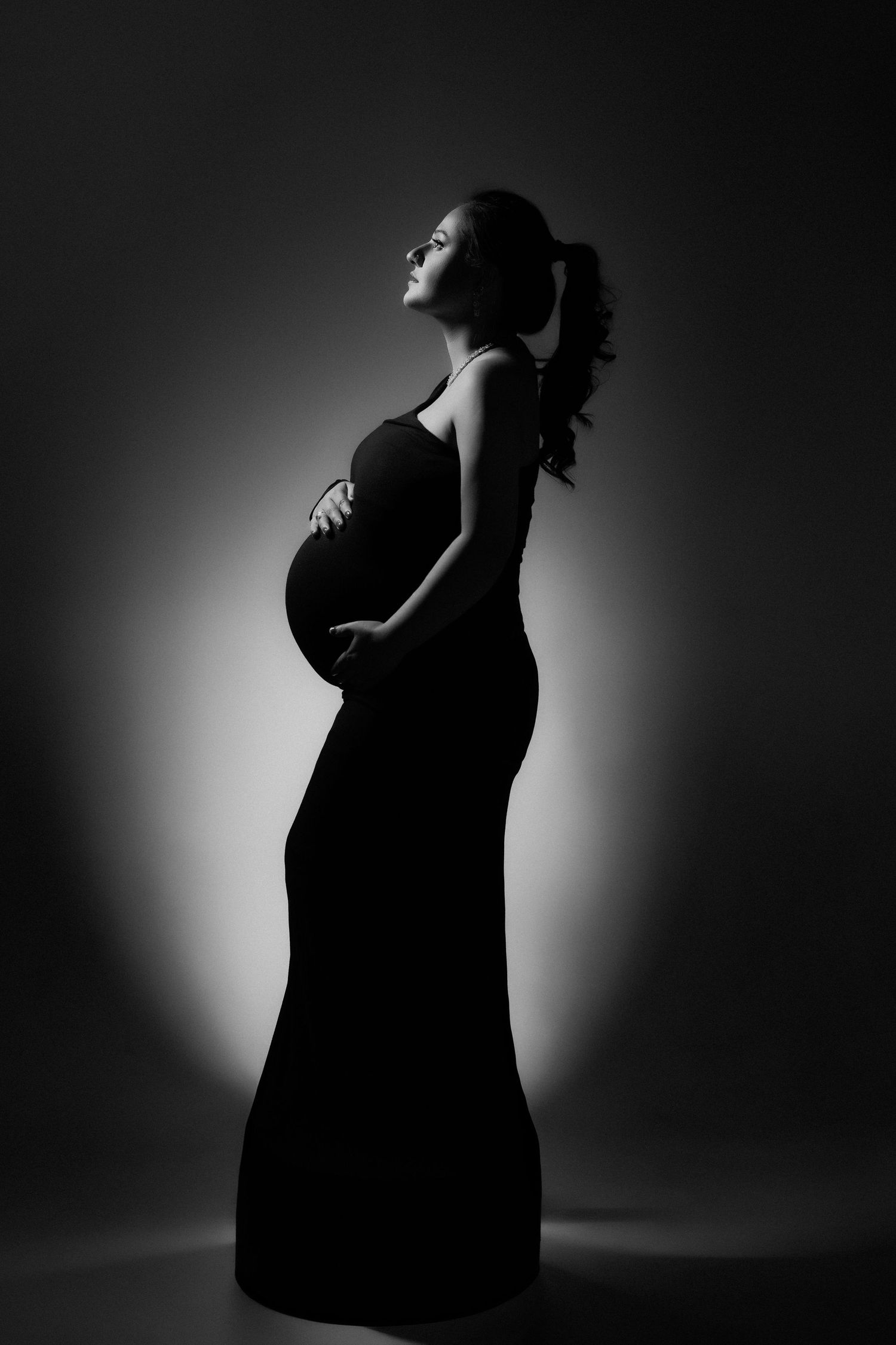 Black and white silhouette of a pregnant woman in a long dress cradling her baby bump, looking upward.