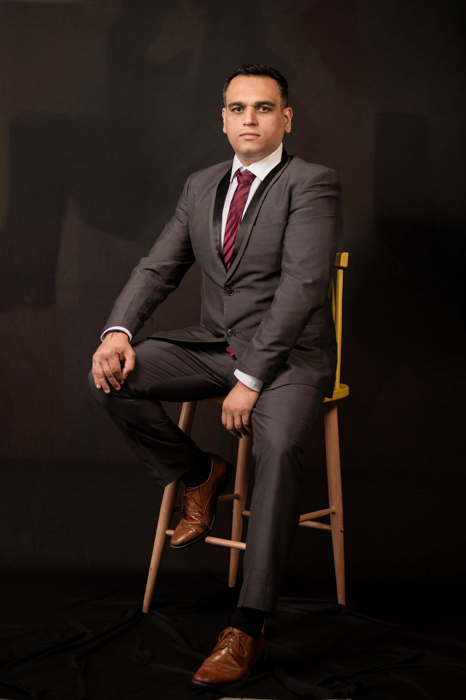 Professional studio portrait of someone in a dark grey suit and burgundy tie seated on a wooden chair against black background.