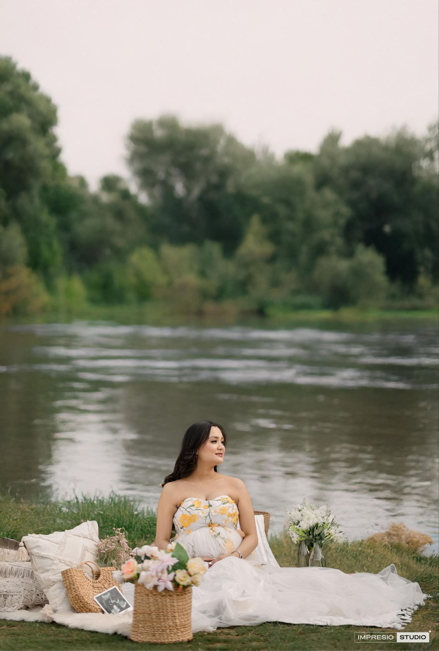 Dreamy riverside maternity photoshoot with picnic setup and flowing white dress.