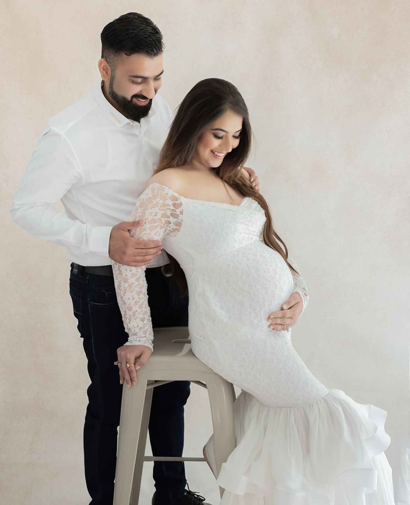 Intimate maternity portrait with couple in white lace gown and casual attire against neutral background.