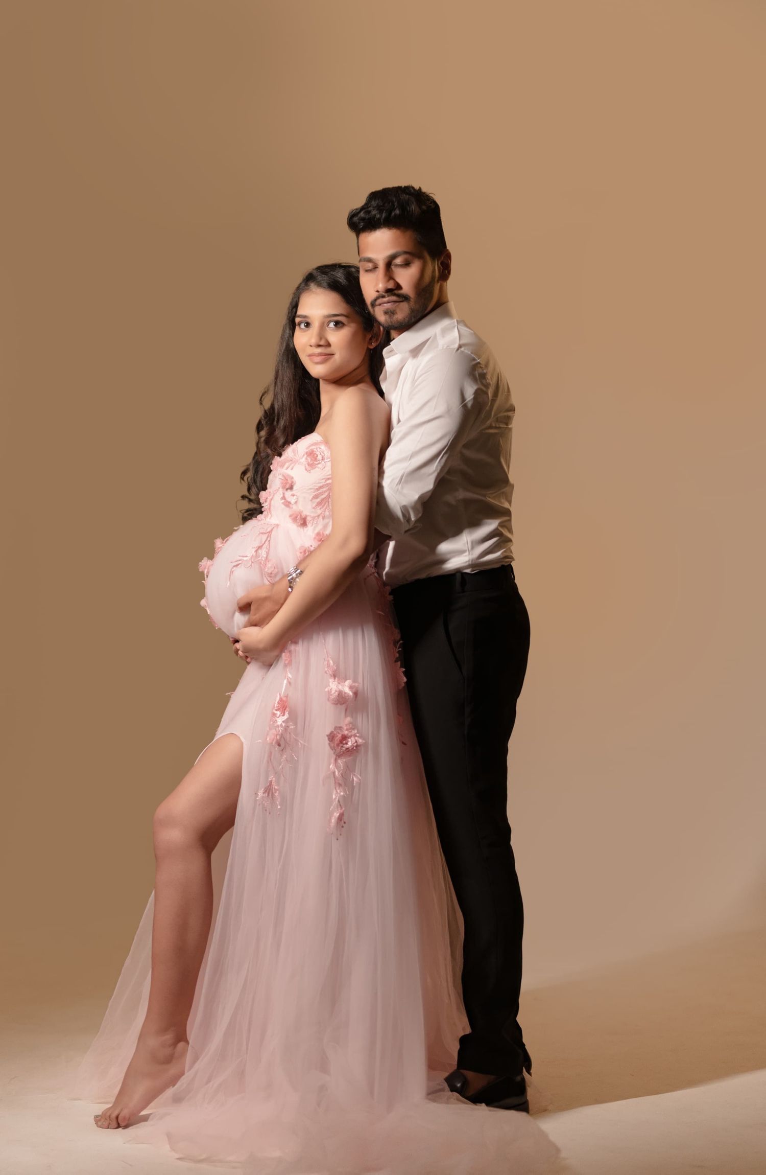 Romantic maternity studio portrait with couple in pink gown and formal attire against brown background.