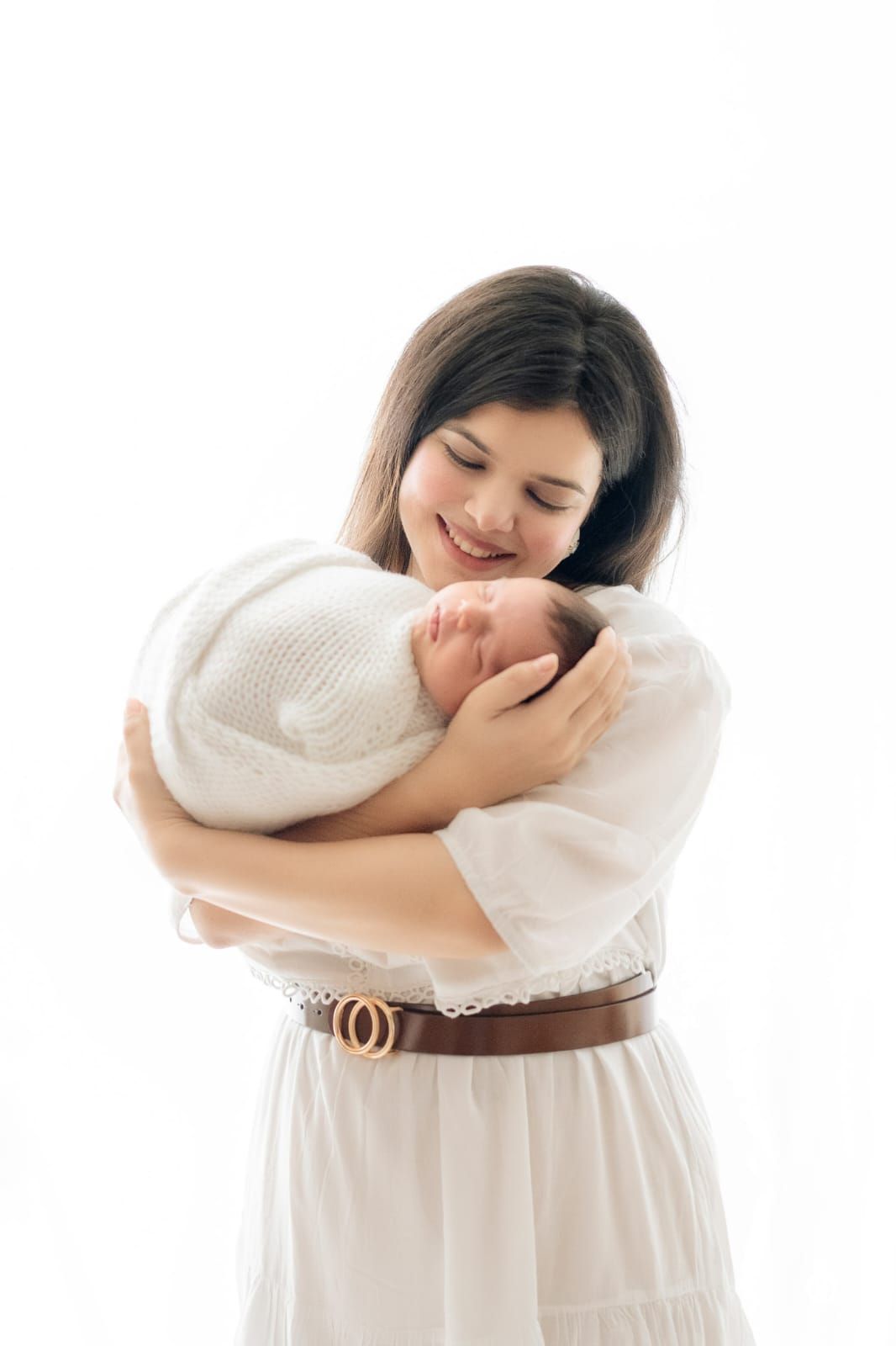 Mother in white dress holding newborn wrapped in soft white blanket against light background.