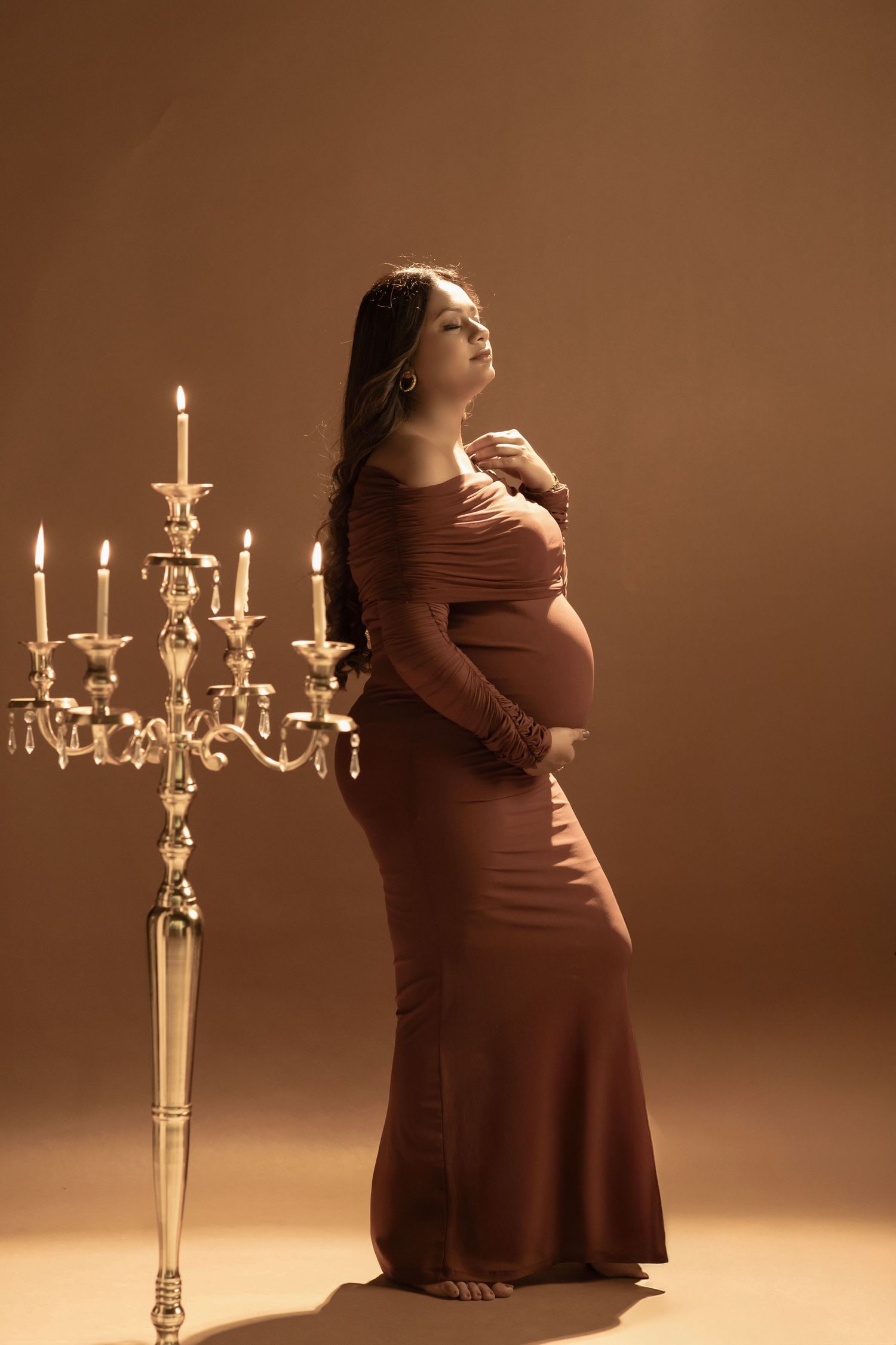 Maternity photoshoot featuring a burgundy dress and elegant gold candelabra against a warm brown background.