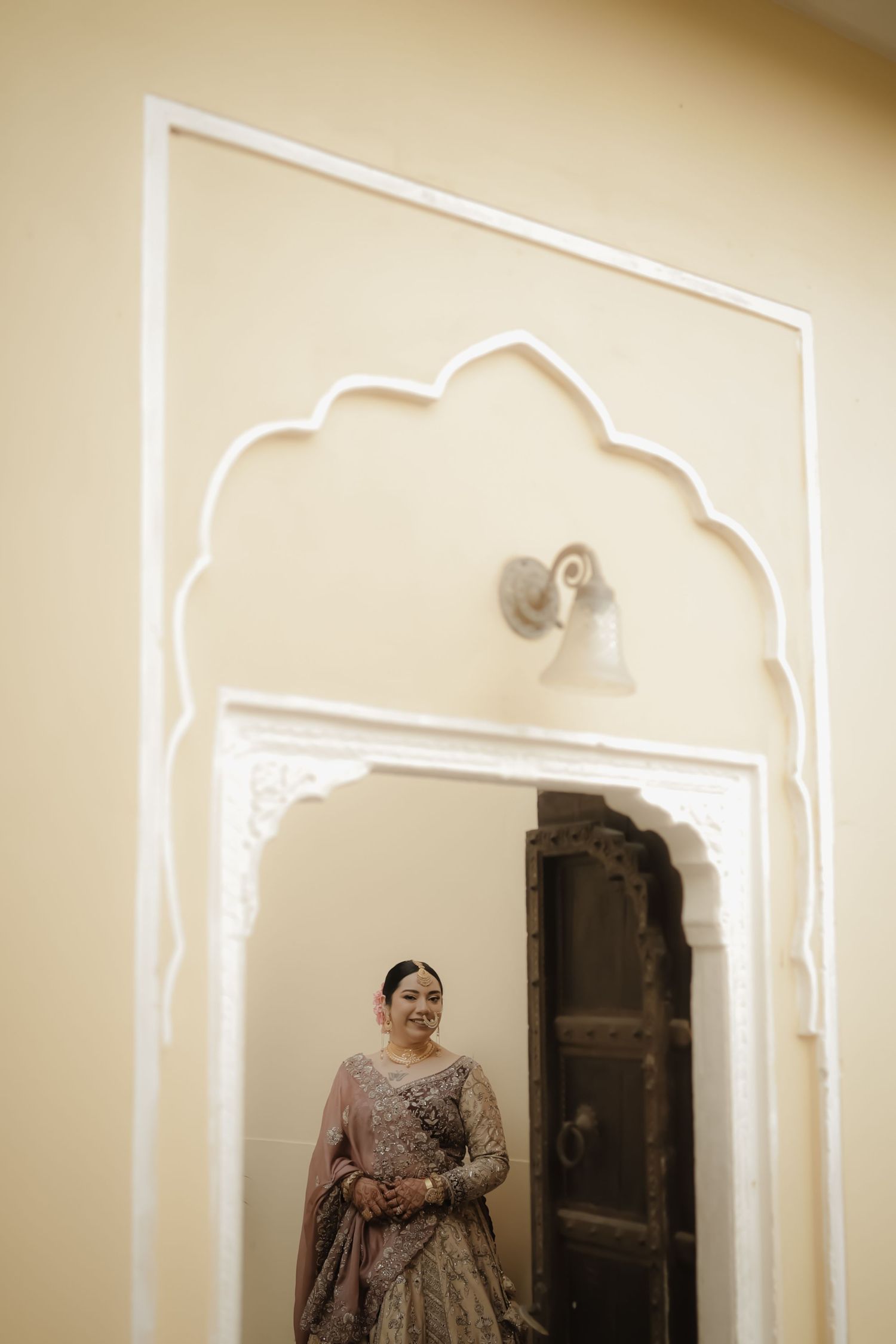 A figure in an ornate beige lehenga stands beneath an elegant scalloped archway in a traditional Indian interior.
