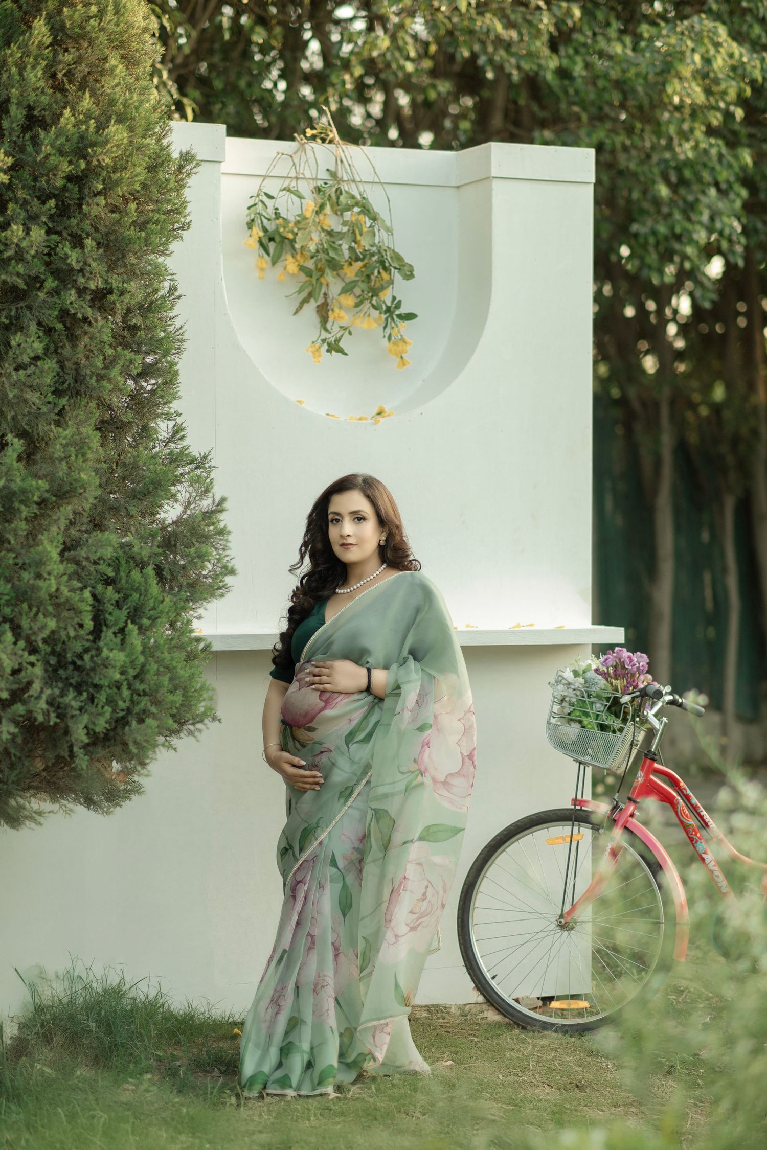 A vintage bicycle leans against a garden wall while someone in a flowing sage green dress poses for maternity photography