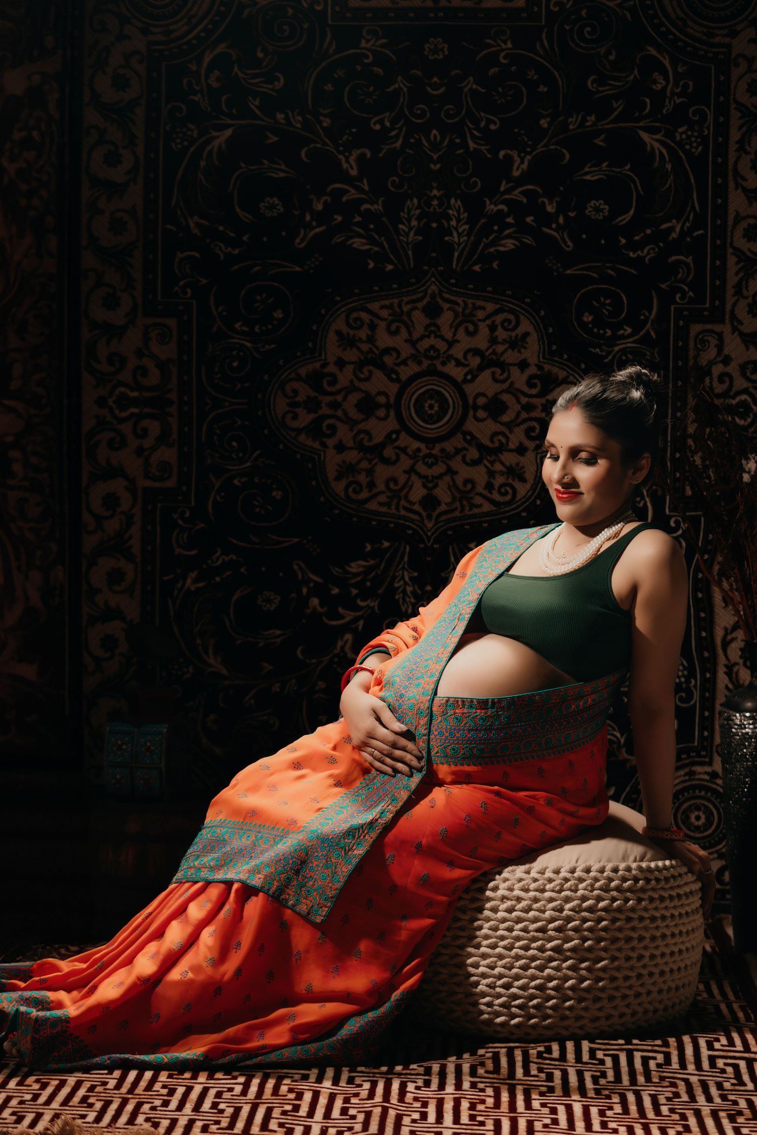 Dramatic maternity portrait against ornate tapestry with orange and gray textiles.