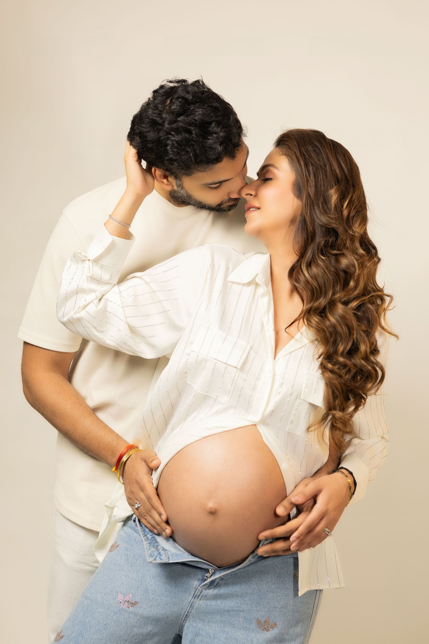 A pregnant couple in matching white shirts share intimate embraces against a beige backdrop in a photo series.