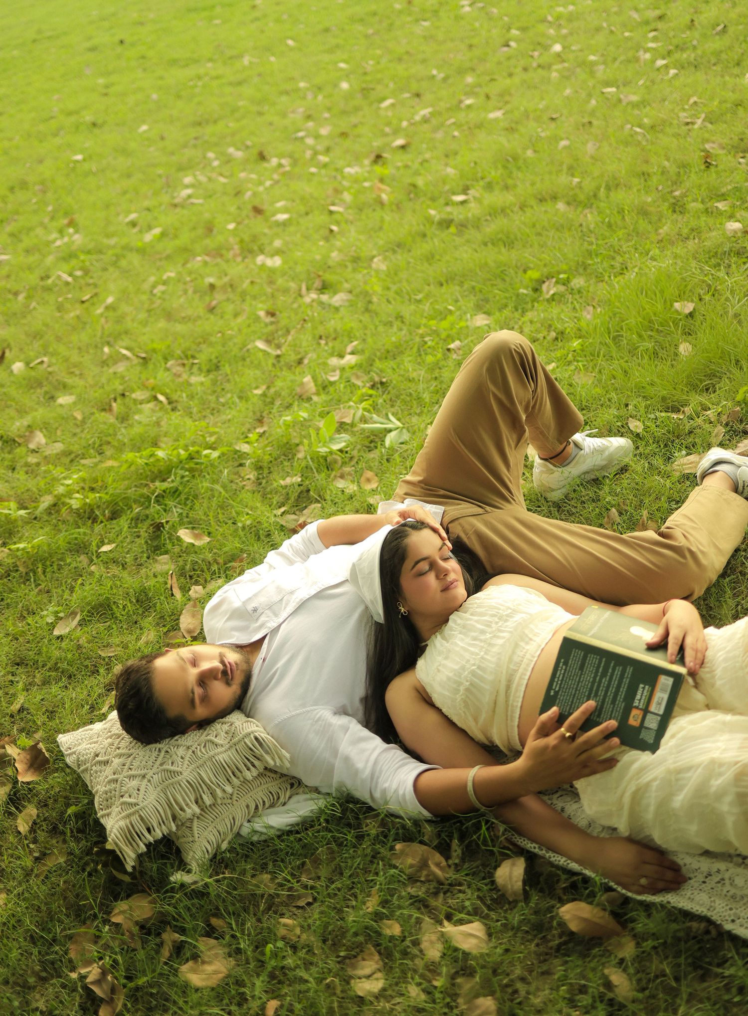 Couple relaxing on green grass in a park, woman resting head on man's chest while holding a book.