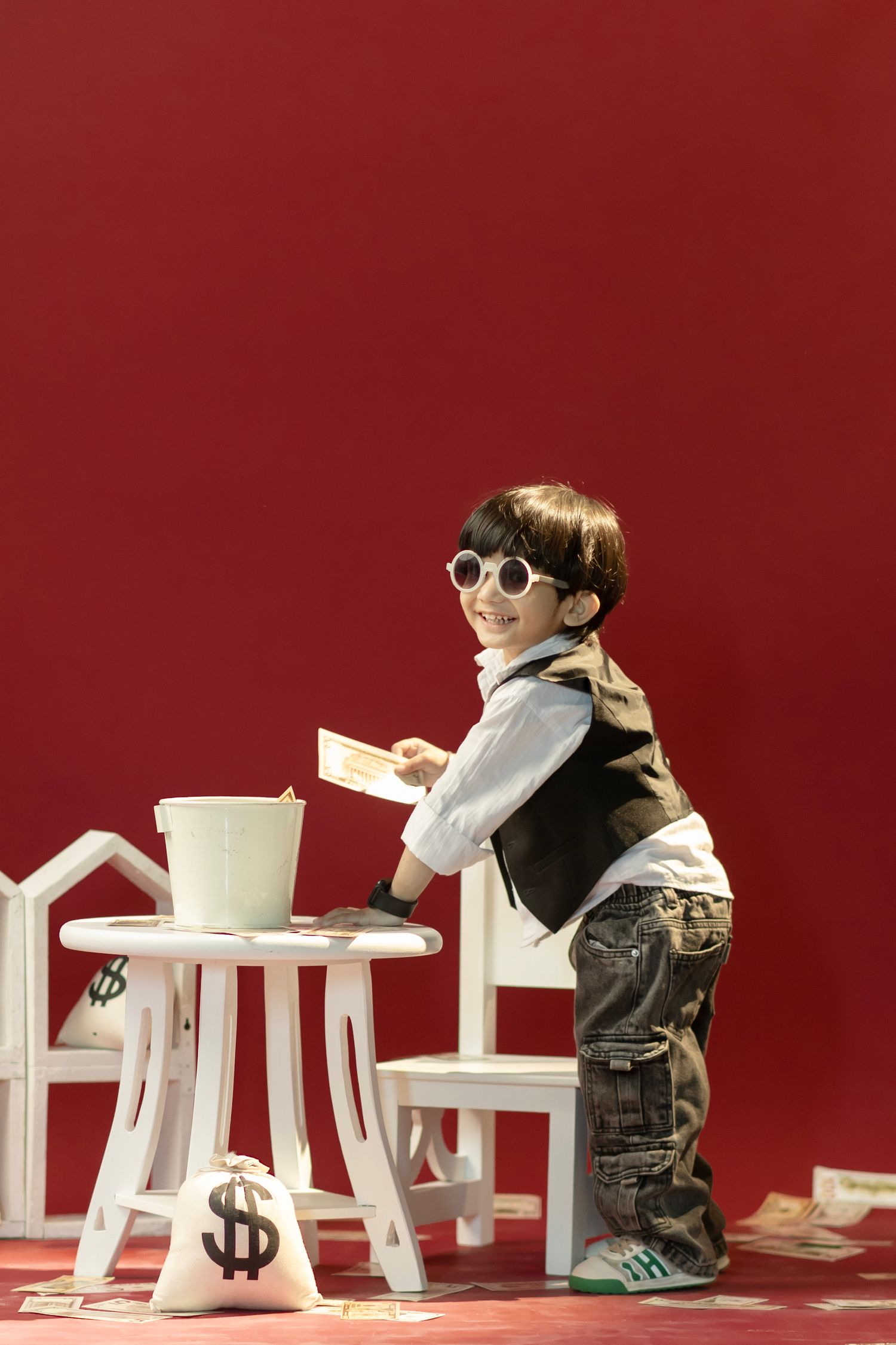Young boy in sunglasses and vest playing with toy money bags and bucket at white table on red background.