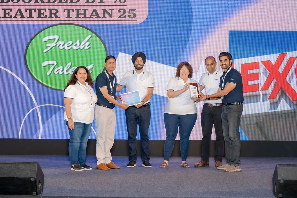Group of people standing on stage at a corporate event with blue backdrop and green circular logos.