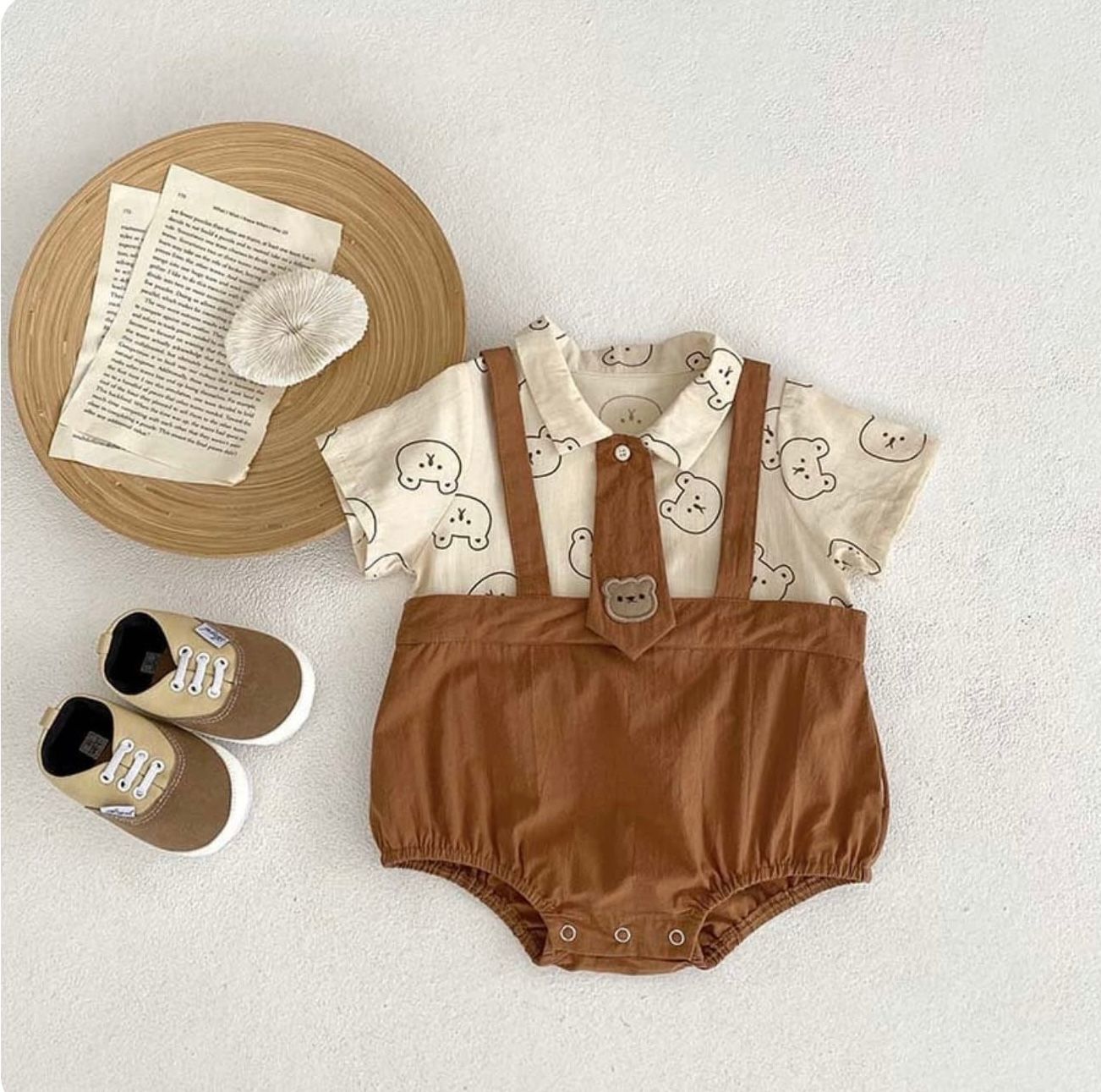 Vintage-style baby outfit with brown suspender bloomers, patterned shirt, beige shoes, and wooden plate display on white surface.