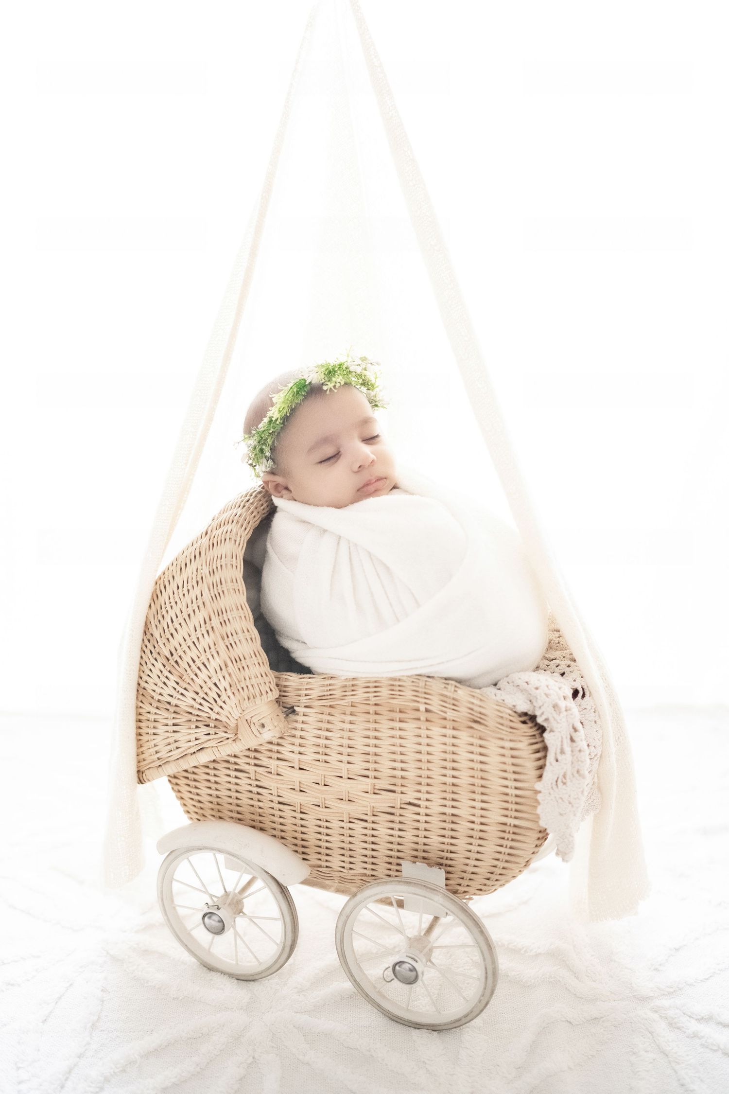 Sleeping newborn baby wrapped in white swaddle with floral crown, resting in a wicker pram on white background.