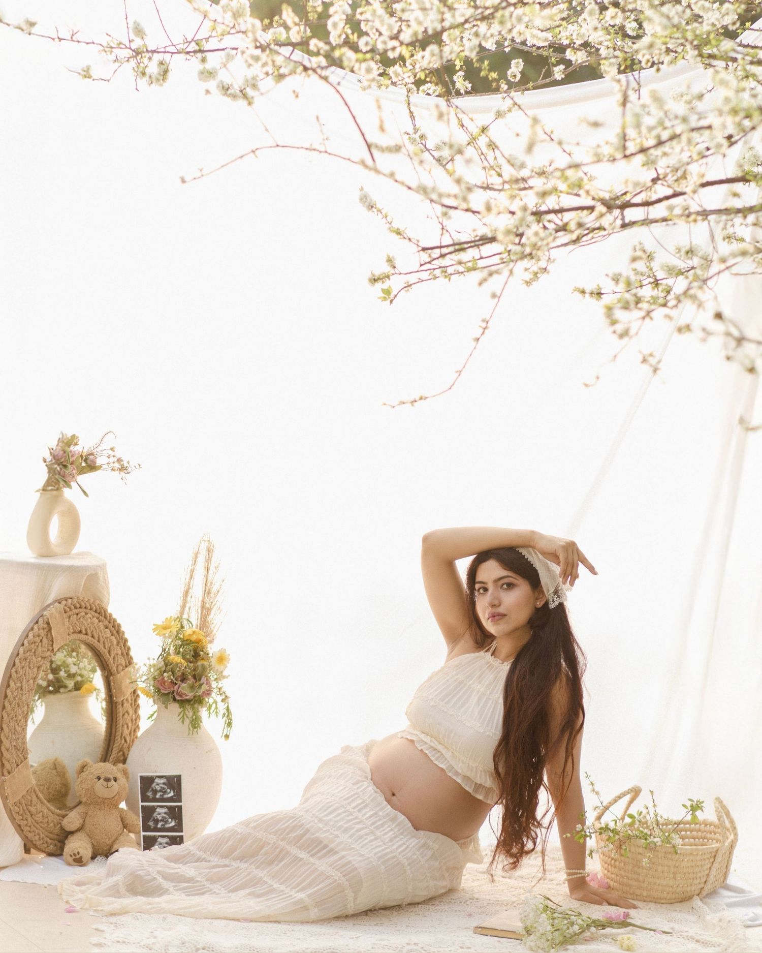 Pregnant woman in white knit crop top and skirt posing gracefully under flowering cherry blossom branches.
