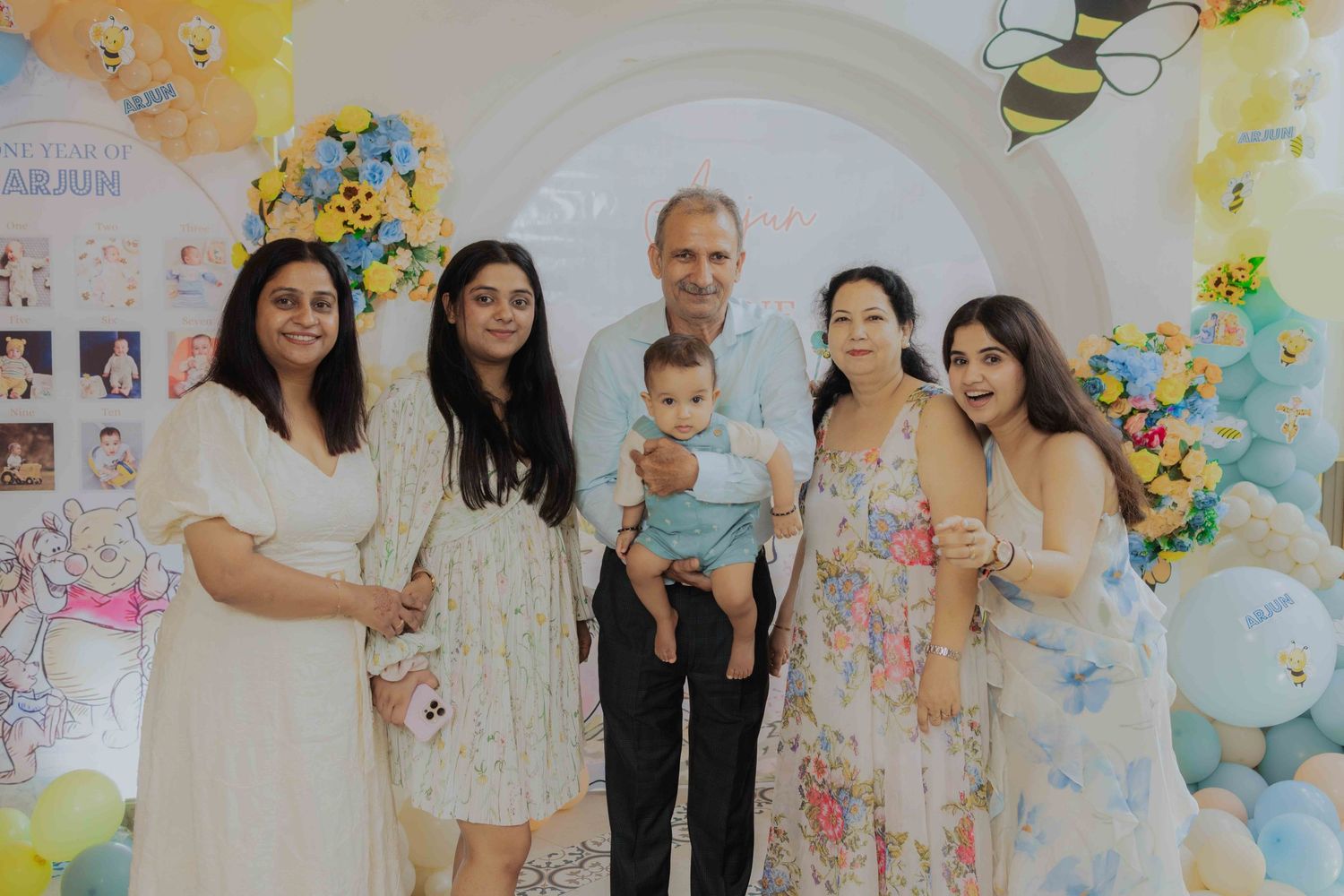 Family group photo at a colorful baby shower with balloon decorations, featuring adults and a baby in festive attire.