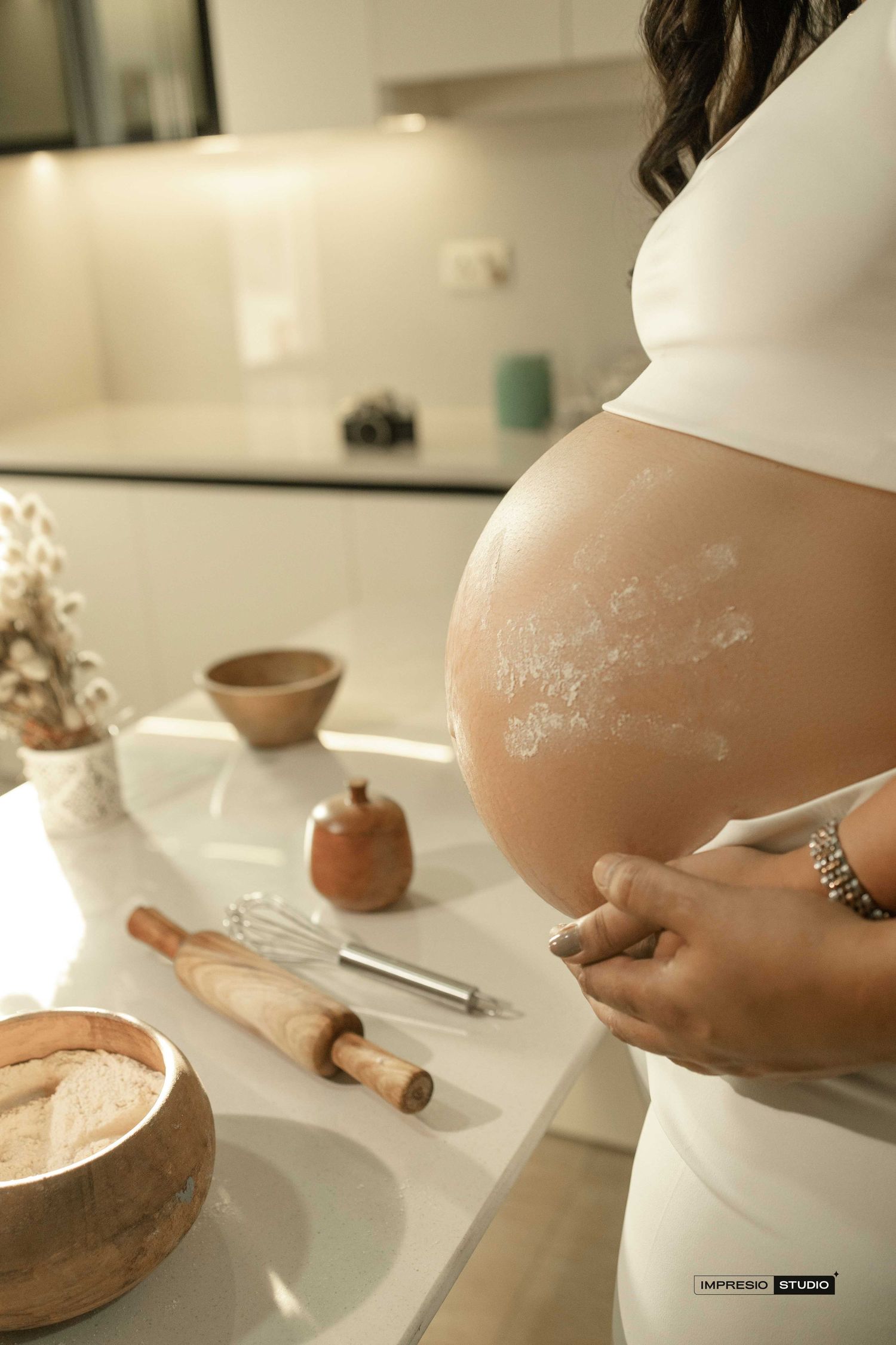 Pregnant woman in white dress with flour on belly baking in modern kitchen with rolling pin and whisk.