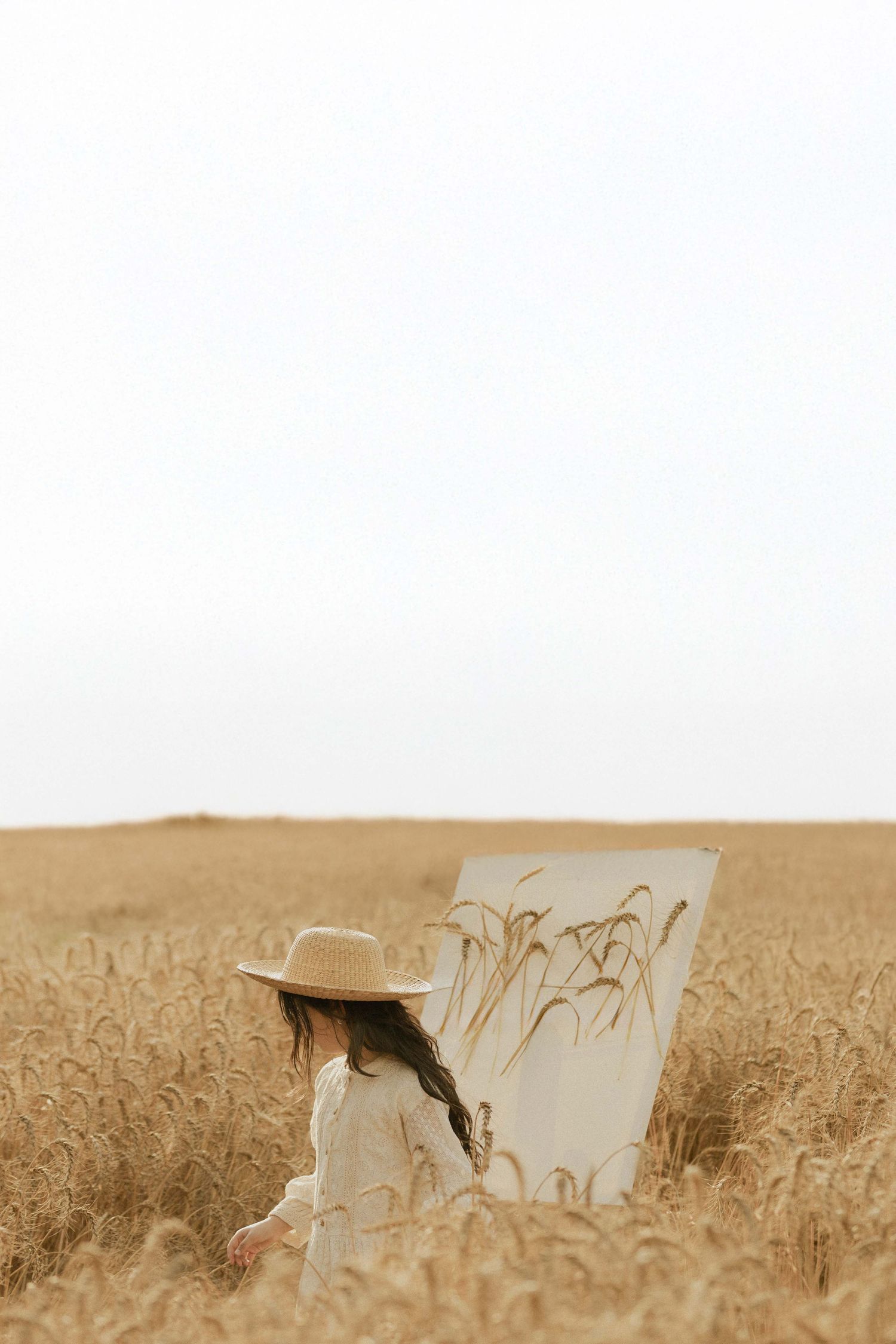A young woman in a straw hat and white dress paints wheat stalks on canvas in a golden wheat field.