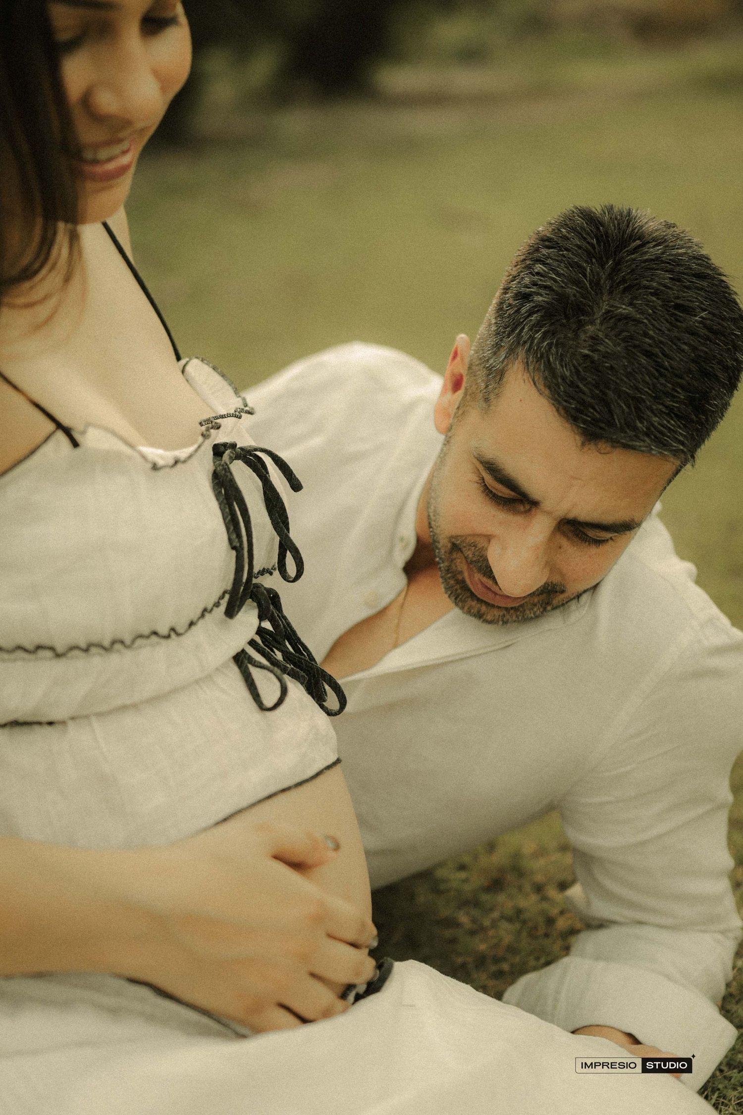 A man tenderly touches a pregnant woman's bare belly during an outdoor maternity photoshoot, both dressed in white.