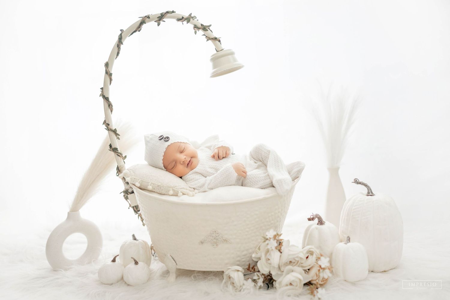 Newborn baby sleeping peacefully in a white basket with floral lamp decor and white pumpkins in a dreamy studio setup.