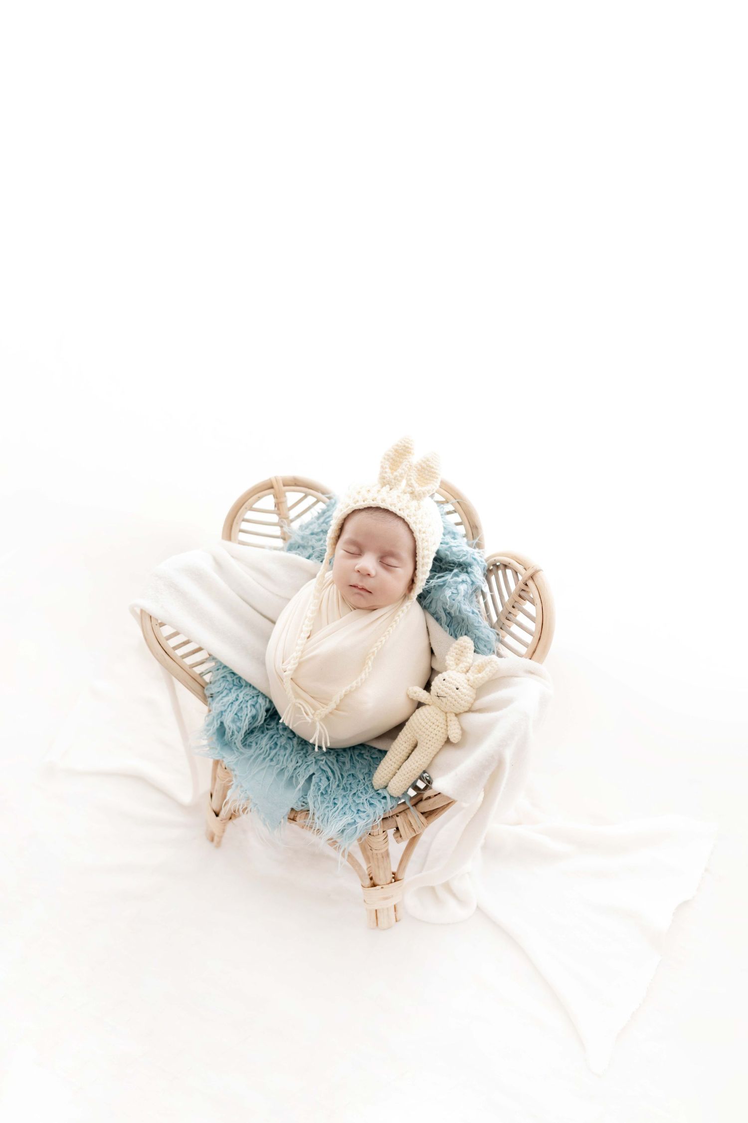 Newborn baby sleeping in a small rattan crib with a blue fur blanket, wearing a cream bunny bonnet with a stuffed toy.