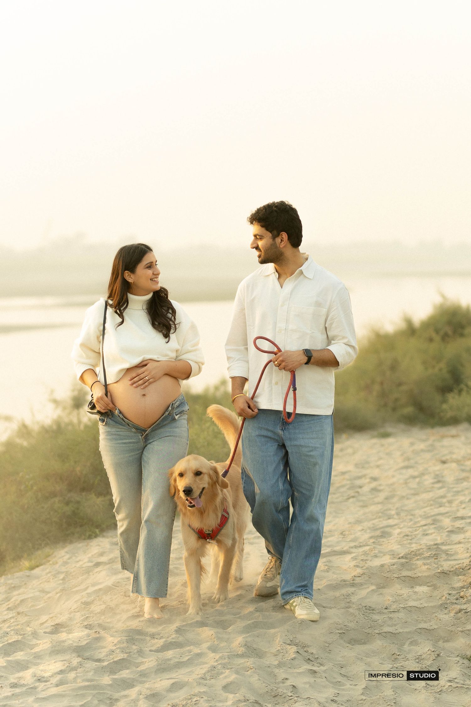 Pregnant woman and man walking golden retriever on sandy beach at sunset during maternity photoshoot.