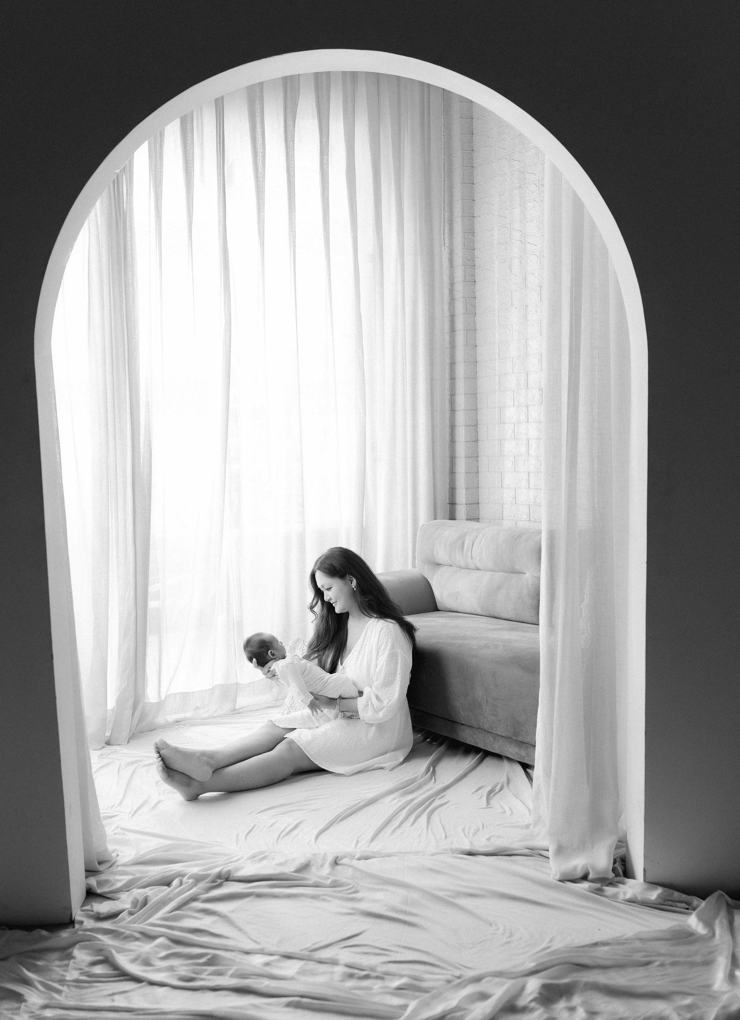 Black and white photo of a mother sitting on the floor holding her newborn baby framed by an elegant arch.