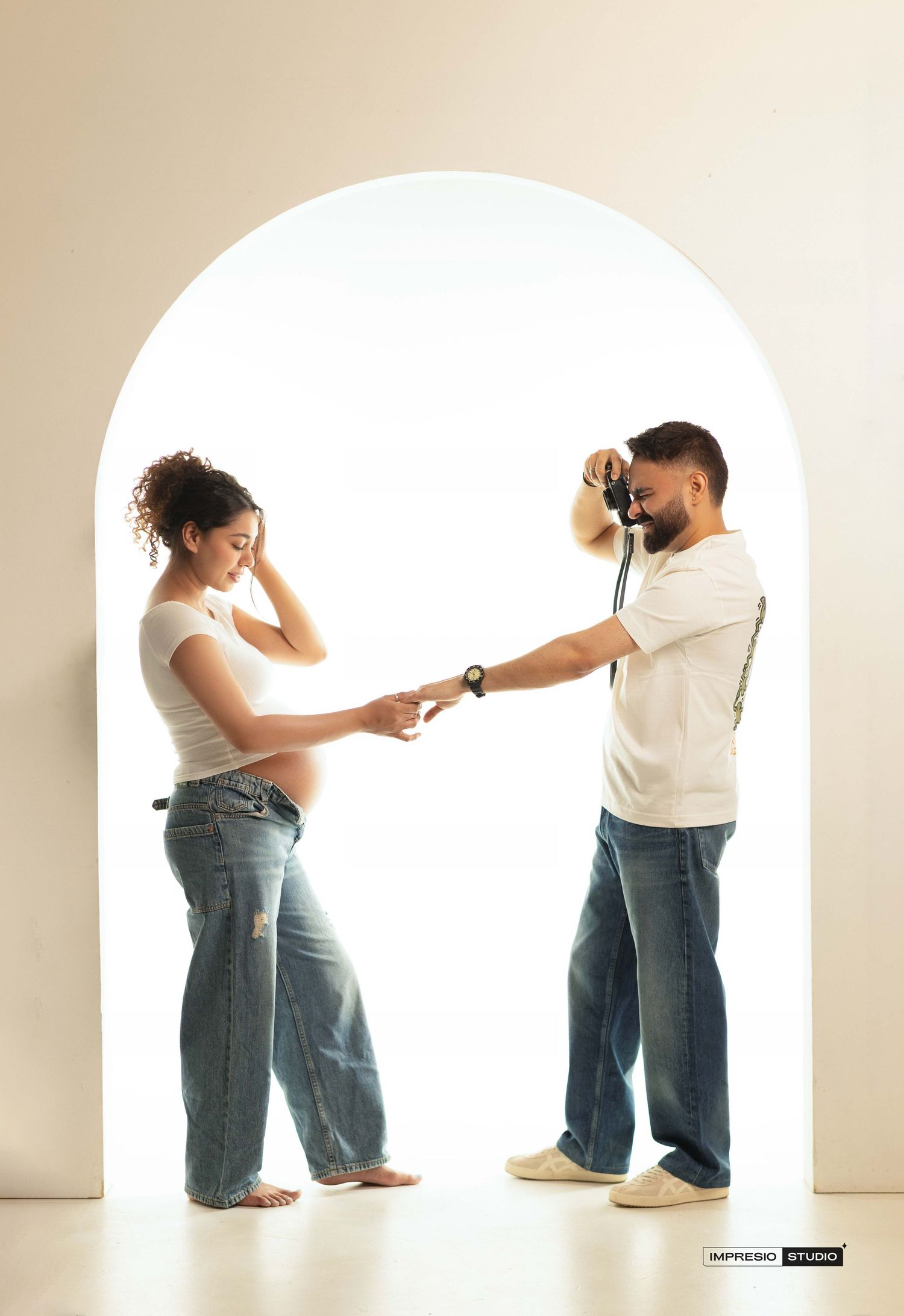 Pregnant woman and man holding hands through a camera strap in an arched doorway, maternity photo shoot.