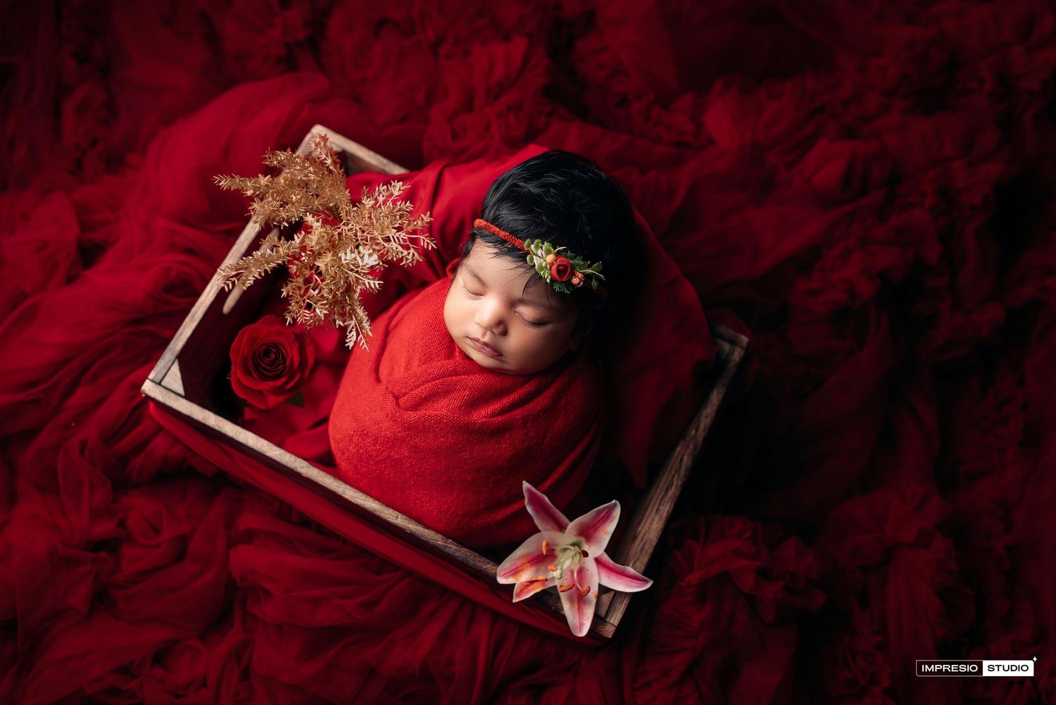 Newborn baby swaddled in red wrap sleeping in wooden frame with red roses and lily flowers on red fabric backdrop.