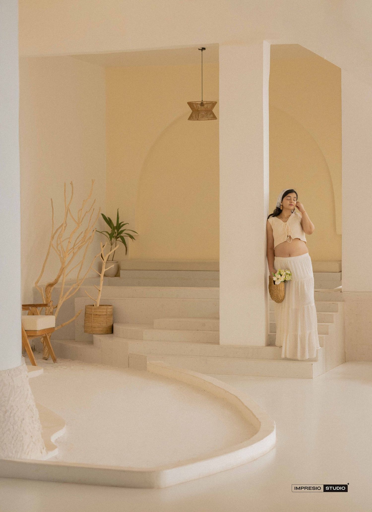 A pregnant woman in a white crop top and skirt holds flowers in a minimalist beige Santorini-style interior.