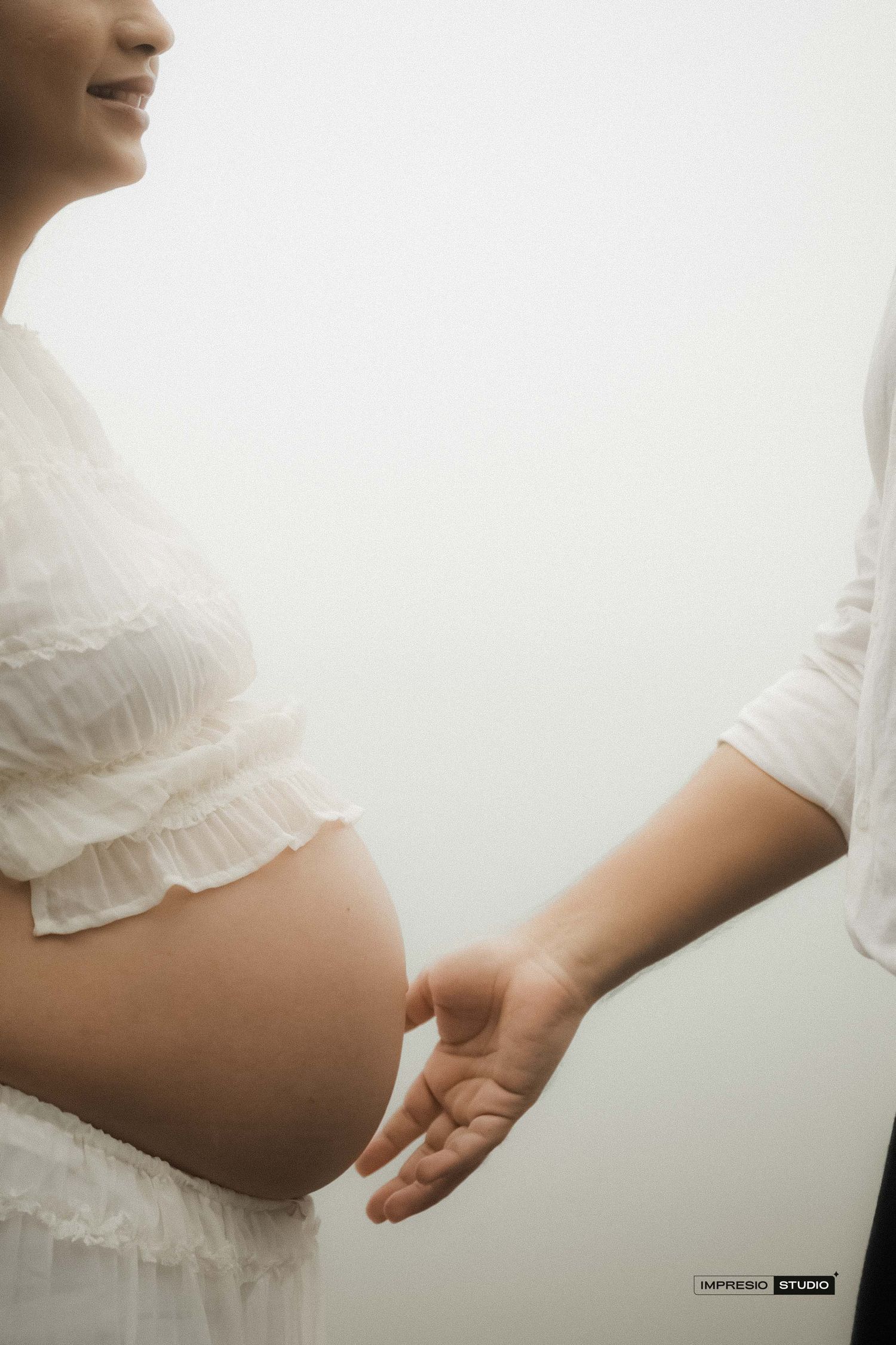 Pregnant woman in white dress with partner holding hands near baby bump in soft light maternity photo.