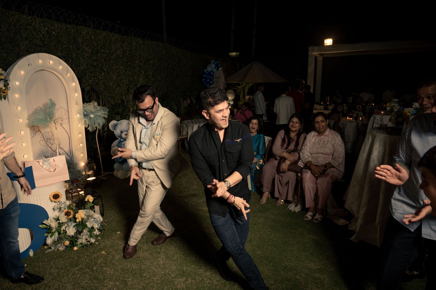 Two men dancing energetically at an outdoor evening wedding celebration with guests seated in the background.