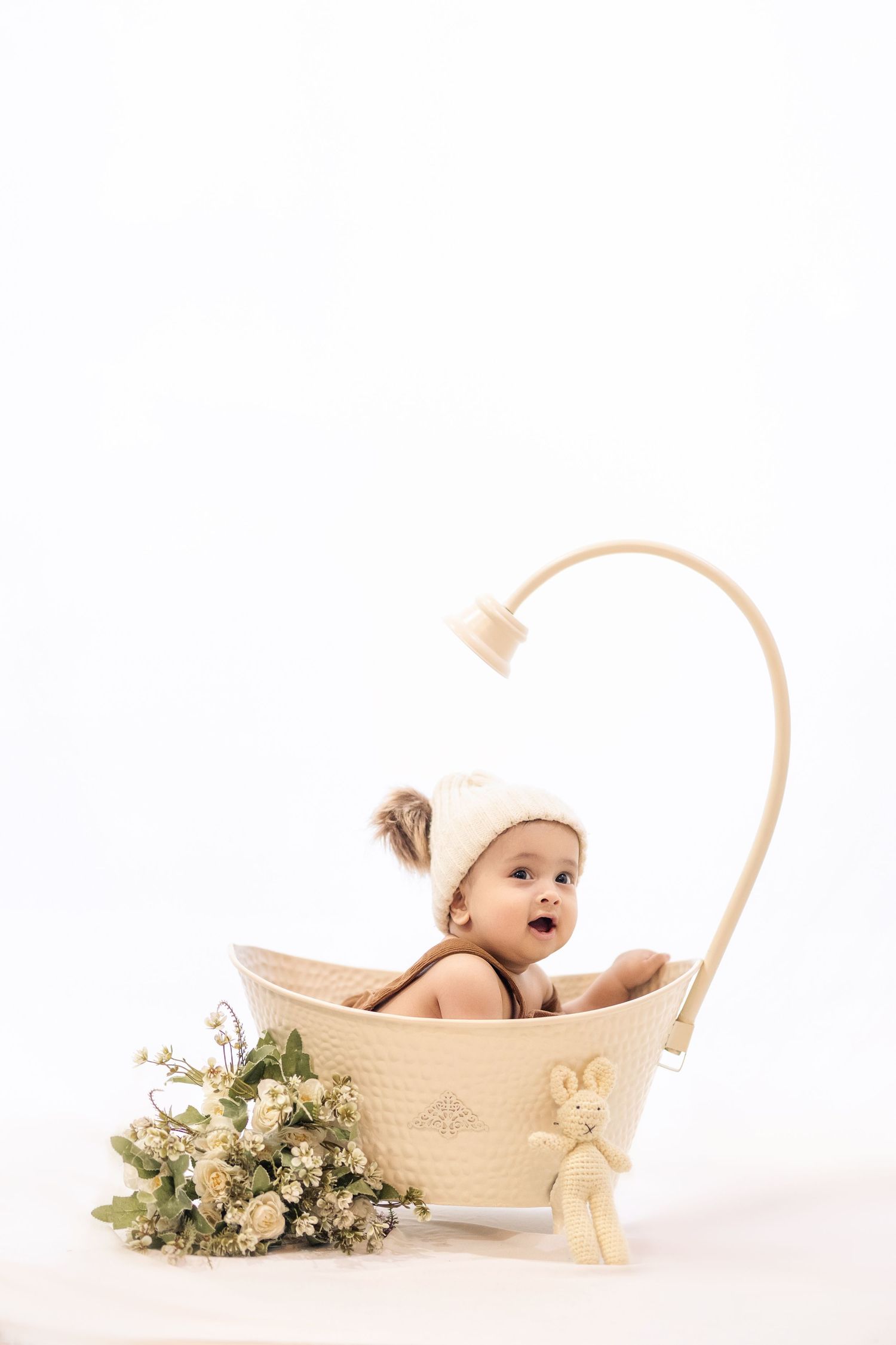 Decorative wooden bathtub prop with shower head contains baby wearing white hat beside floral arrangement.