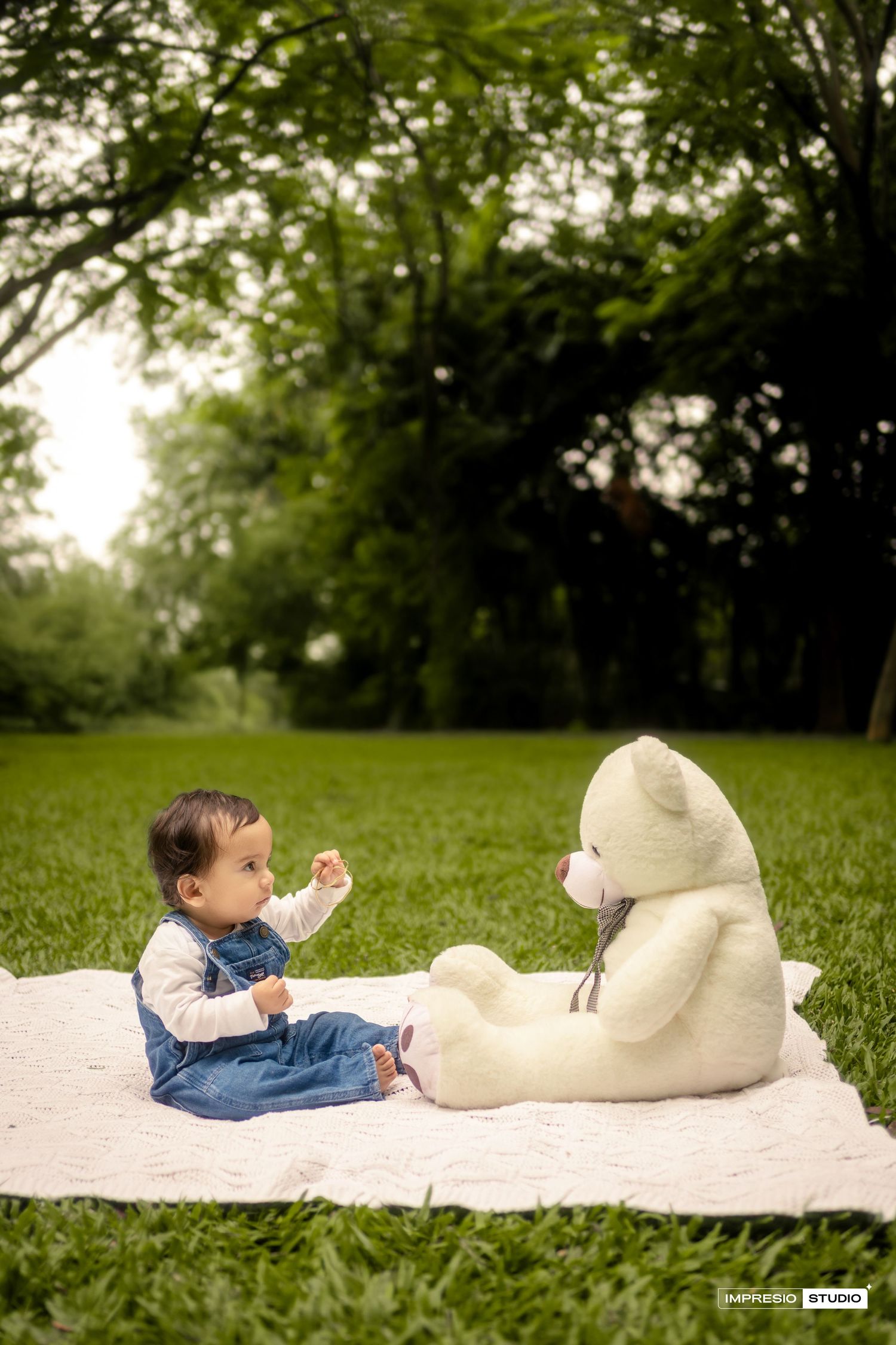 A baby in denim overalls sits on a white blanket in a grassy park with a large teddy bear.
