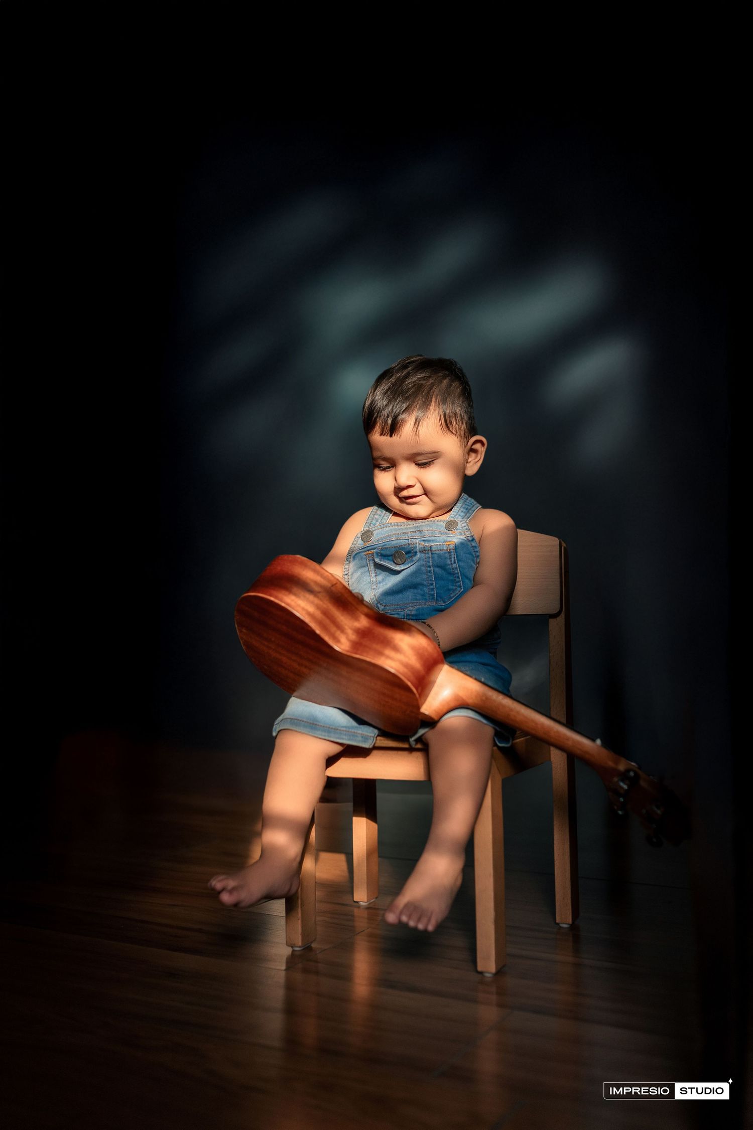 A small child in overalls sits barefoot on a wooden chair holding a large acoustic guitar in a dramatic studio setting.