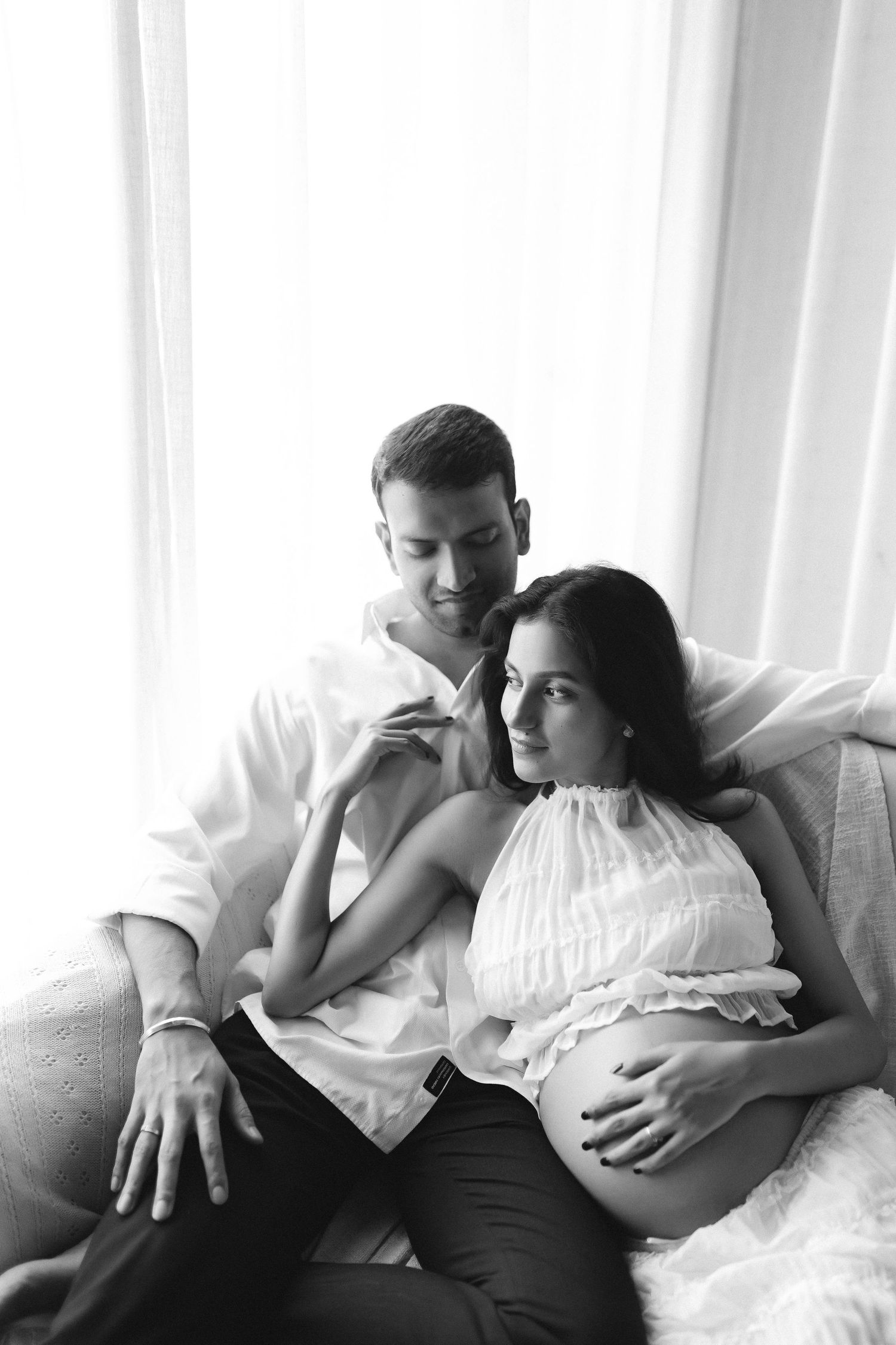 A black and white intimate maternity photo of a couple relaxing together on a couch by a window.
