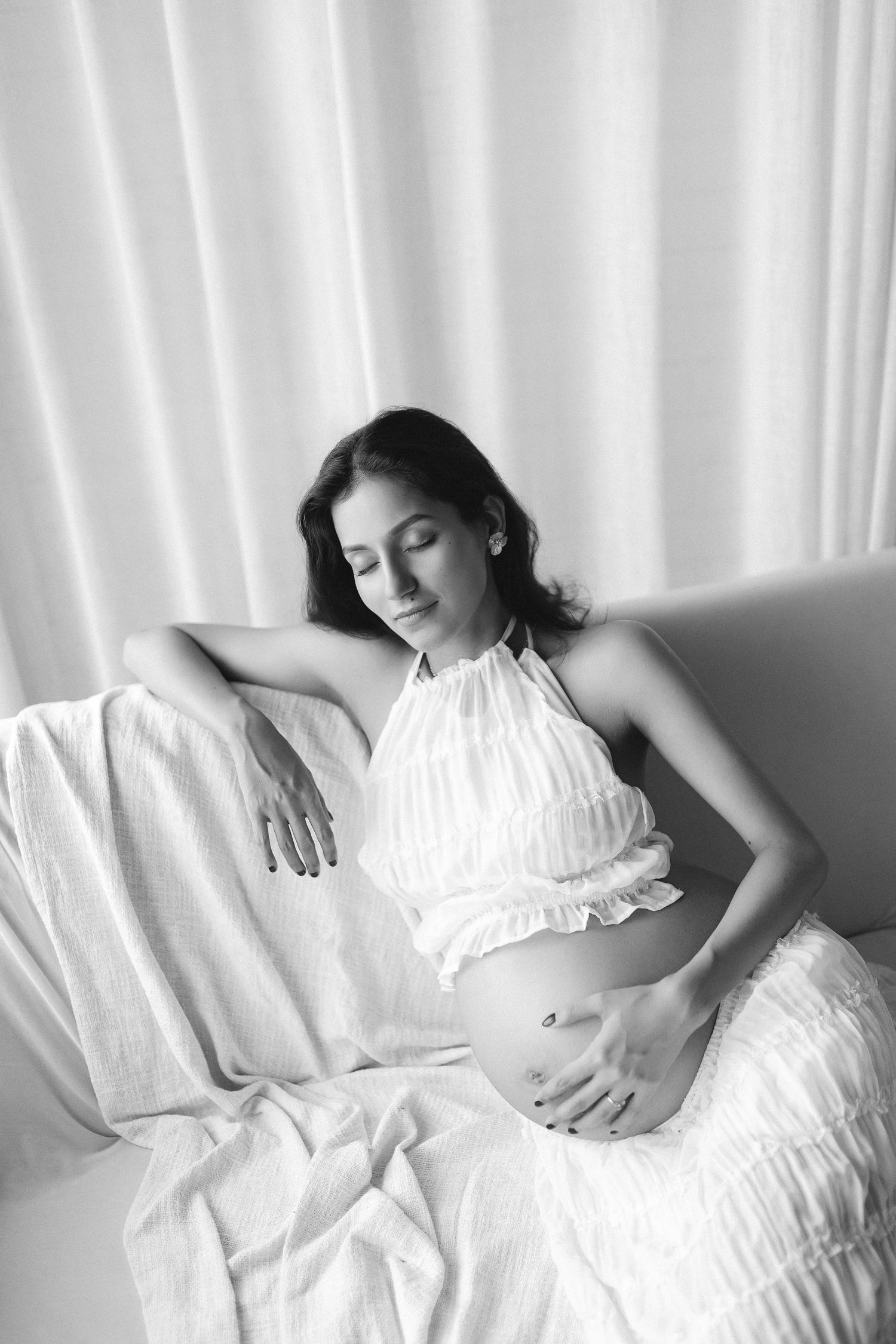 A black and white portrait of someone in a white ruffled dress reclining on white bedding in soft window light.