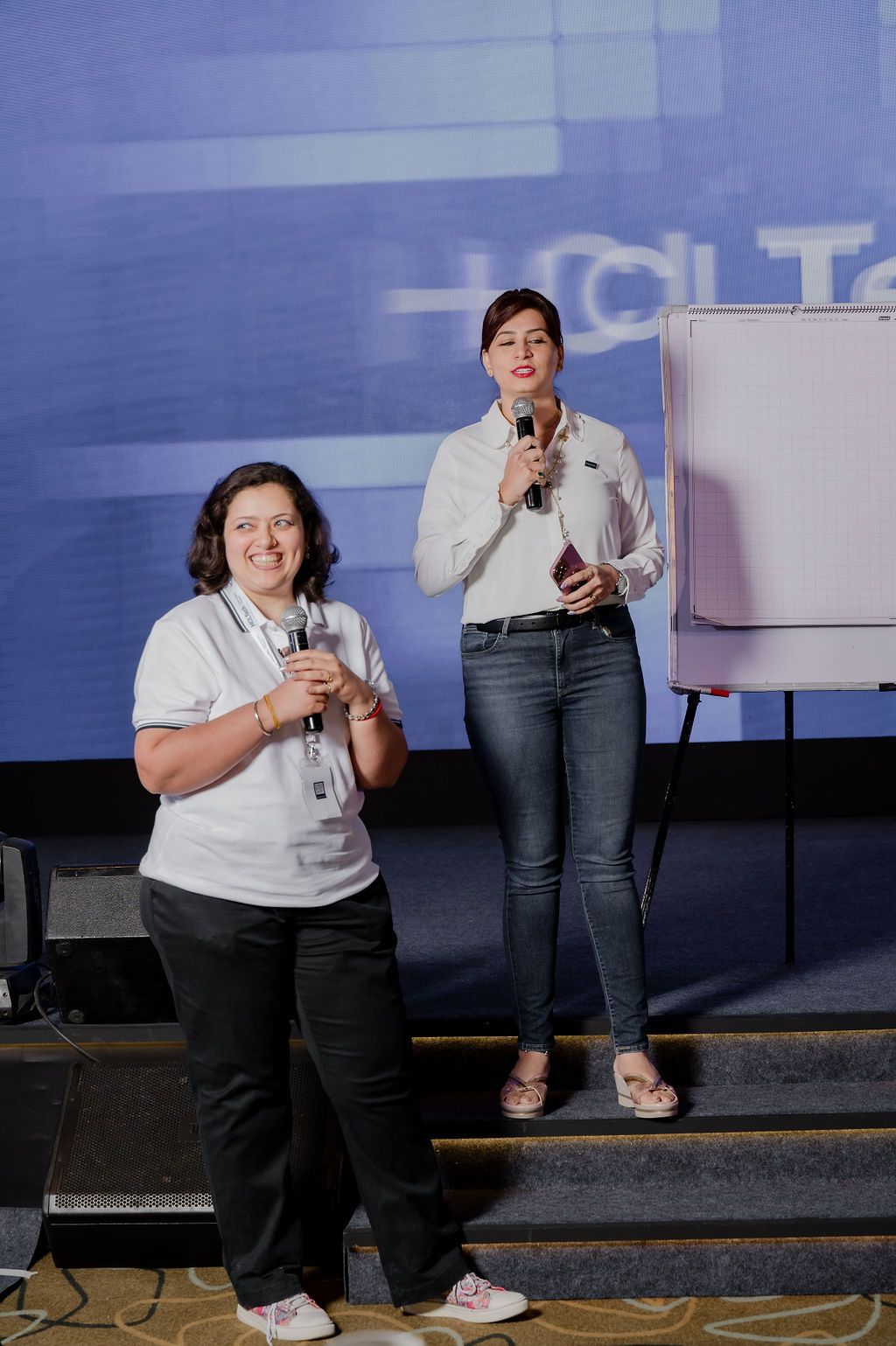 Two presenters stand on stage with microphones during a professional conference presentation.
