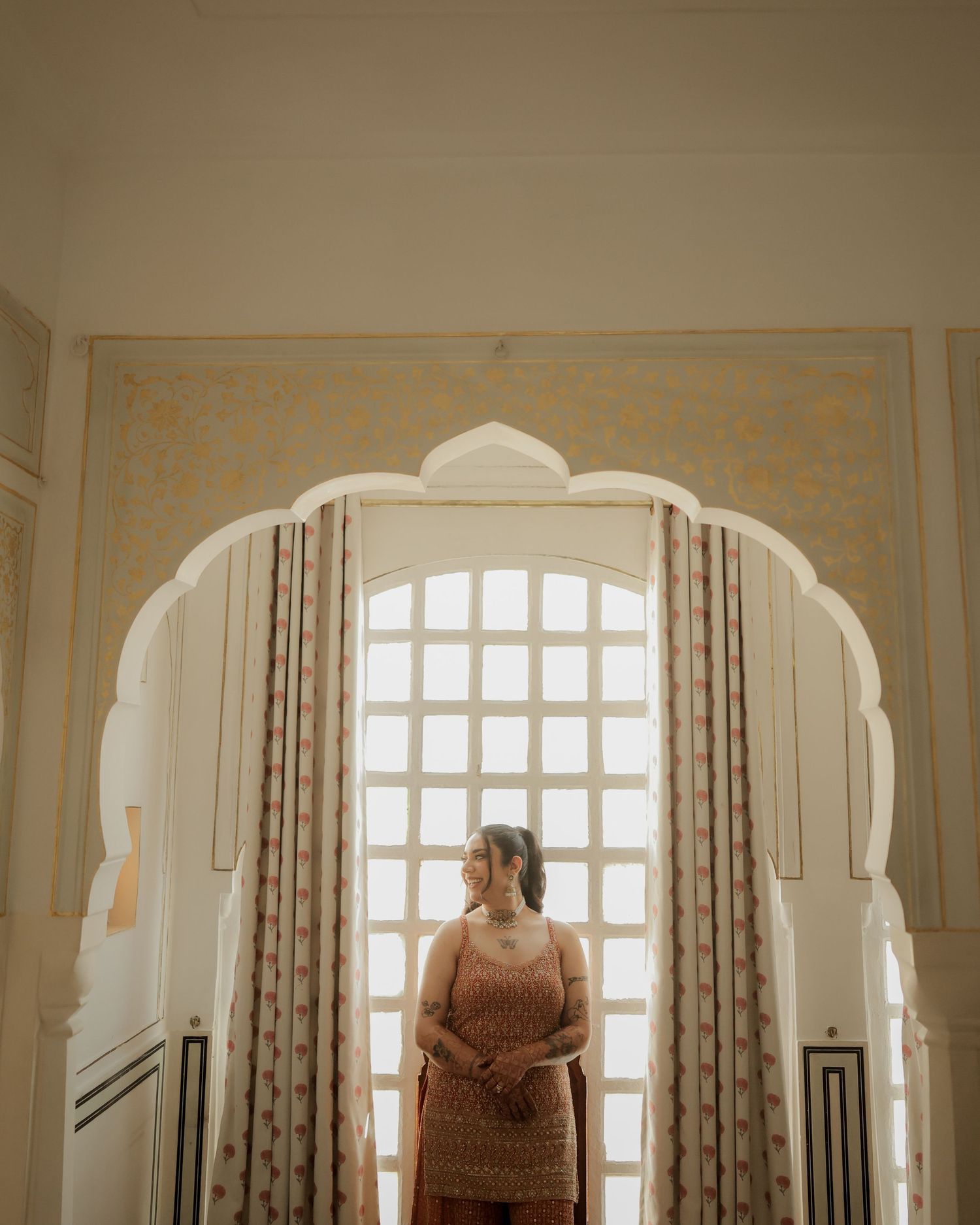 Woman in traditional Indian attire standing under ornate white arch with latticed window backdrop.