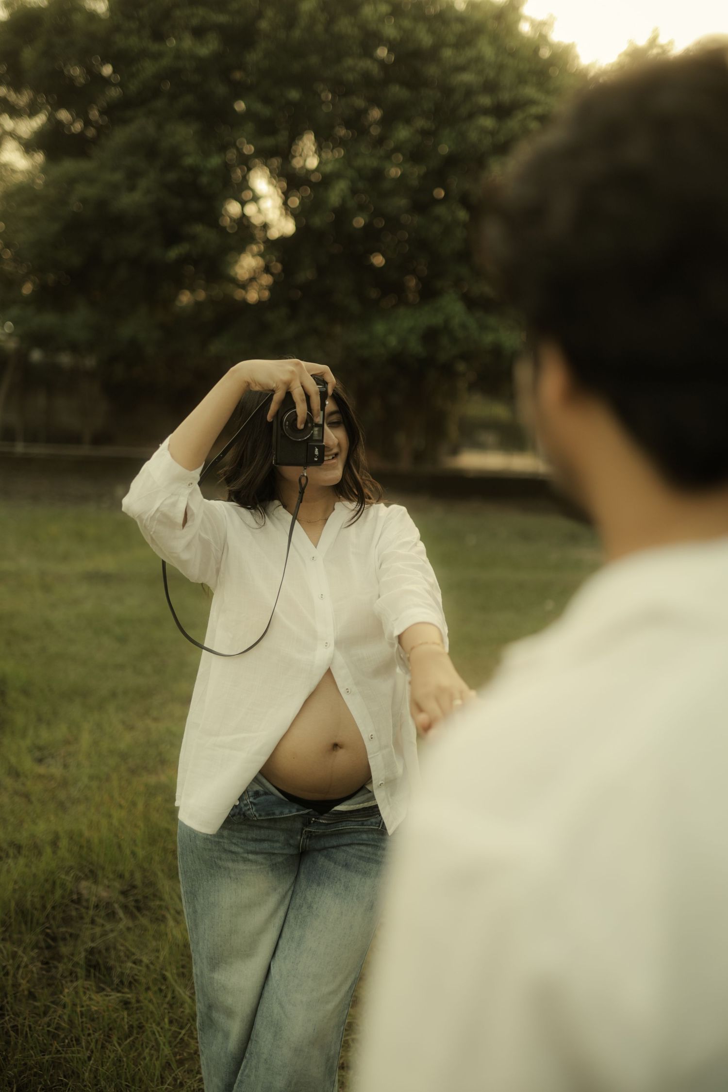 Pregnant woman in white shirt and jeans photographs partner outdoors in lush green park setting.