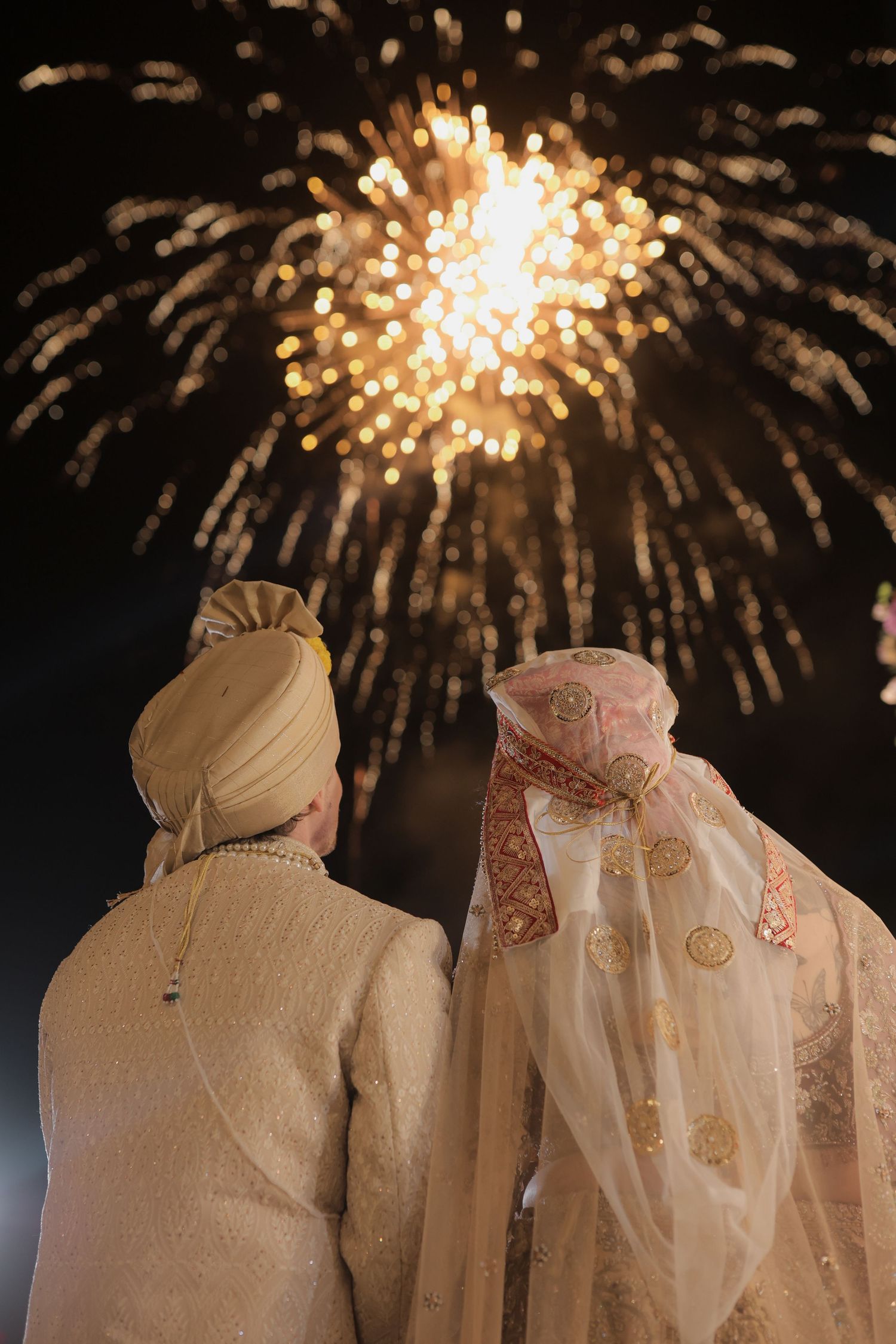 A couple in traditional wedding attire watches fireworks display in the night sky during their celebration.