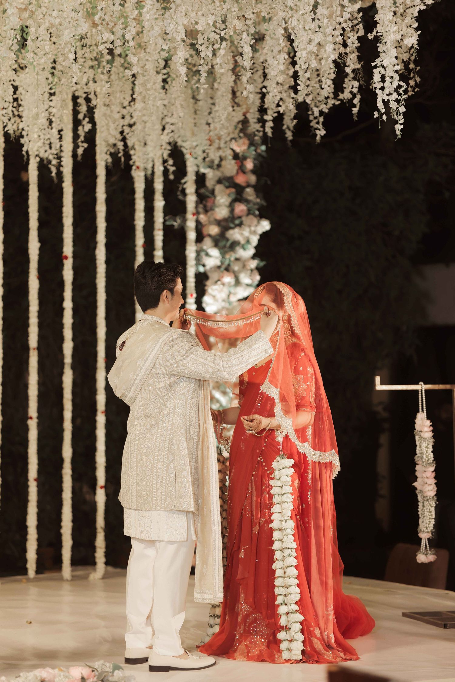 Groom placing red dupatta on bride in traditional Indian wedding ceremony under white floral canopy.