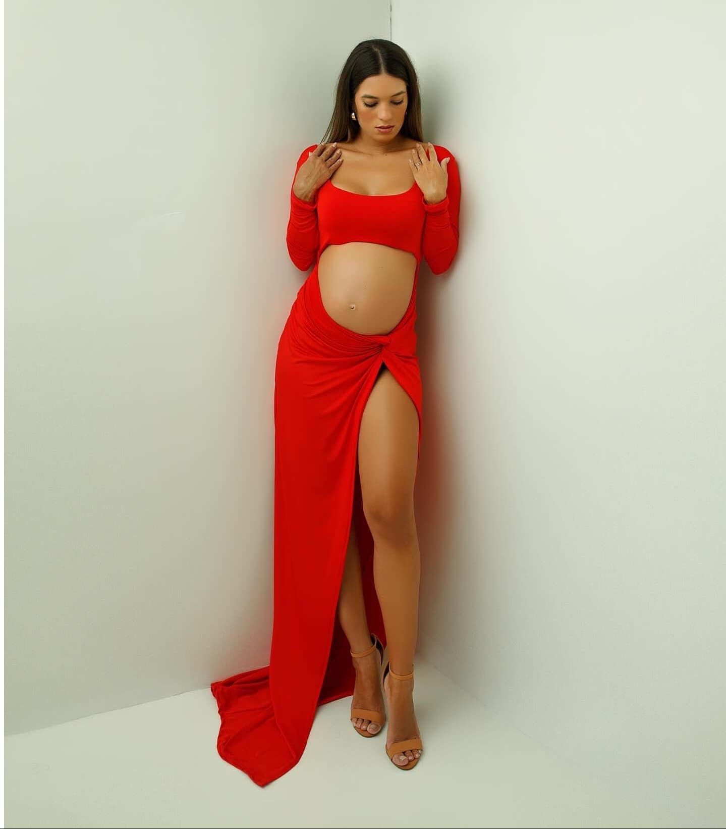 Elegant red maternity gown with cutout design and high slit photographed against white wall.