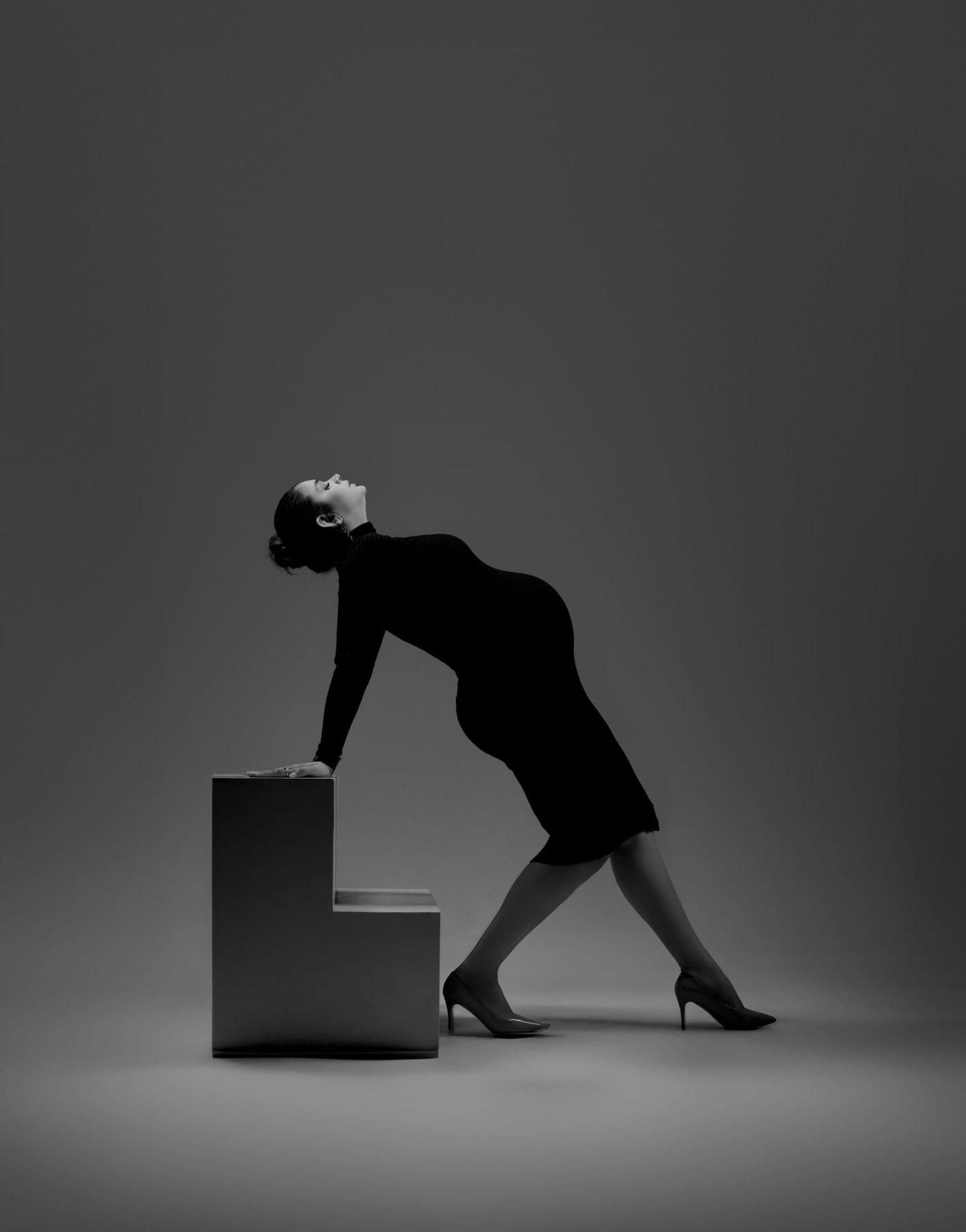 Dramatic black and white photo of pregnant woman in dress leaning back on cube in artistic pose.