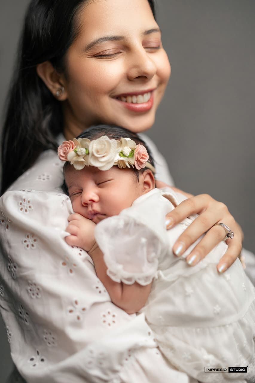 Smiling mother in white dress holds sleeping newborn baby wearing a floral crown, studio portrait photography.