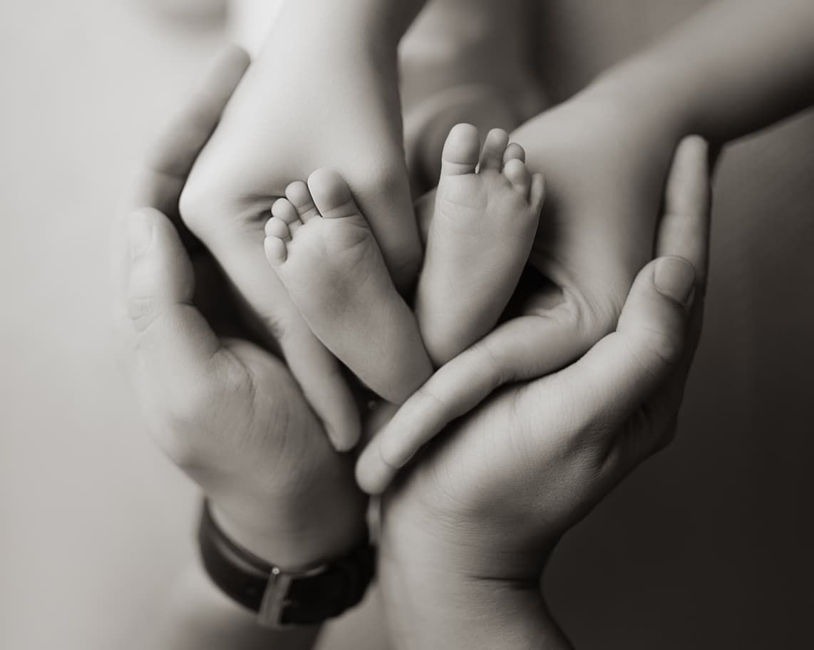 Two parents cradle their newborn baby's tiny feet in their hands in black and white.