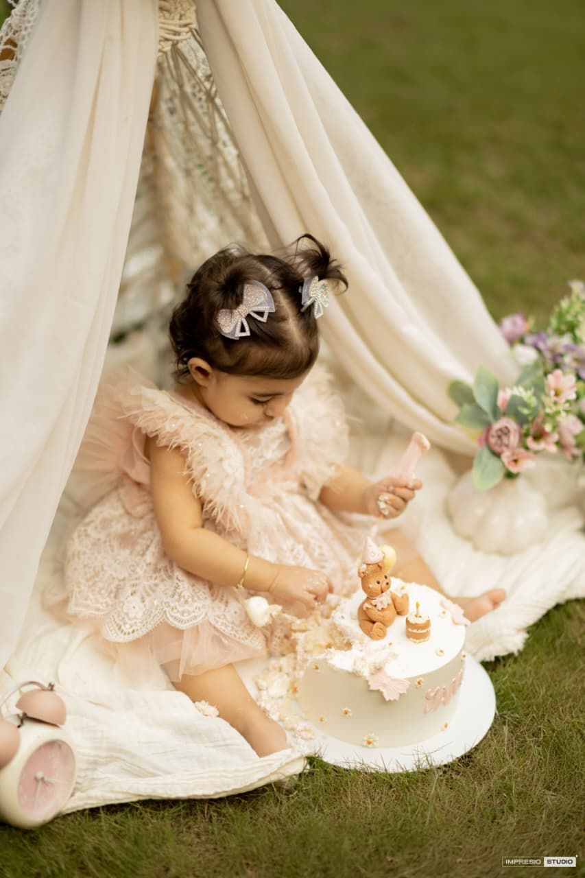 Toddler girl in pink lace dress smashing her first birthday cake under a boho teepee tent outdoors.