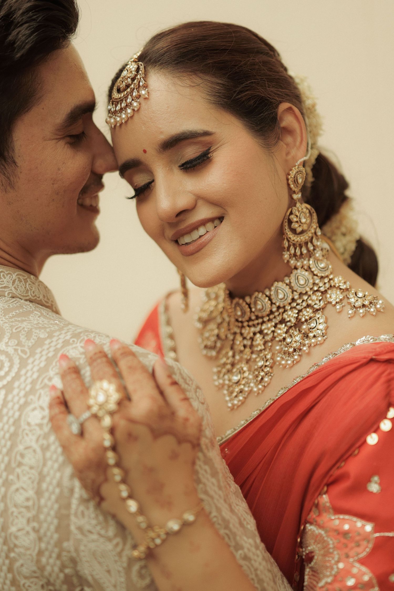Indian bride in red saree with gold kundan jewelry smiles closely with groom in white sherwani.