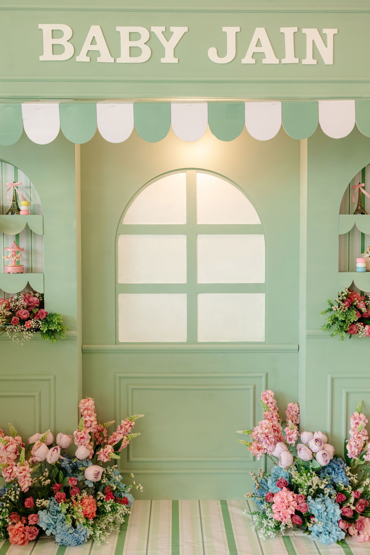 Mint green Baby Jain baby shower backdrop with floral arrangements, arched window, and striped awning decoration.