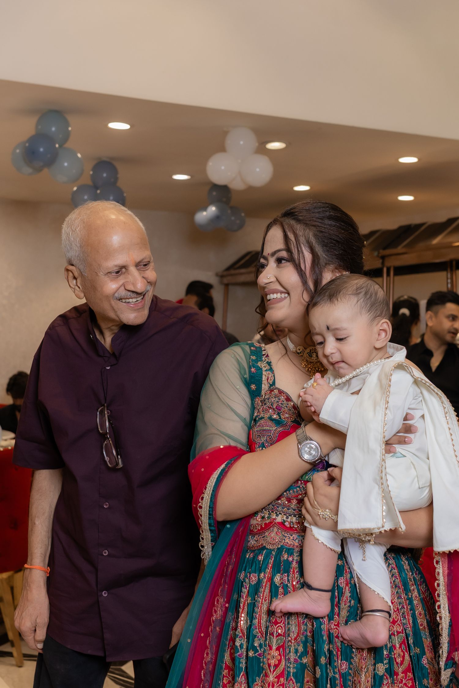 Smiling woman in a colorful embroidered outfit holds a baby beside a joyful older man at a festive indoor party with balloons.