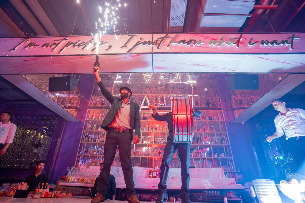 Men celebrating in a vibrant nightclub with sparklers, colorful lights, and a well-stocked bar in the background.