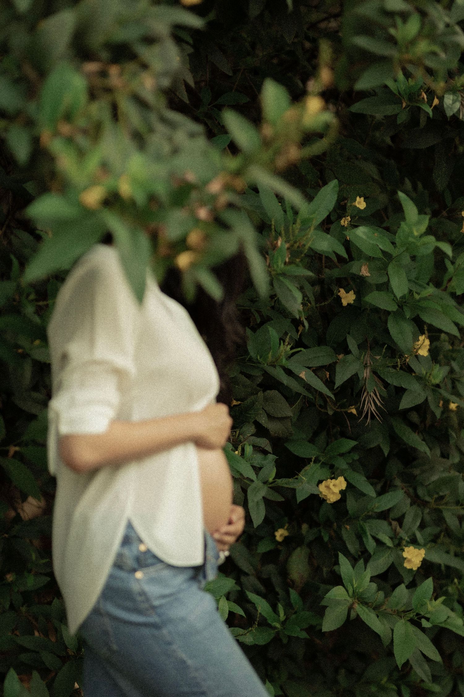 Pregnant person in white sweater and jeans standing among lush green bushes with small yellow flowers.